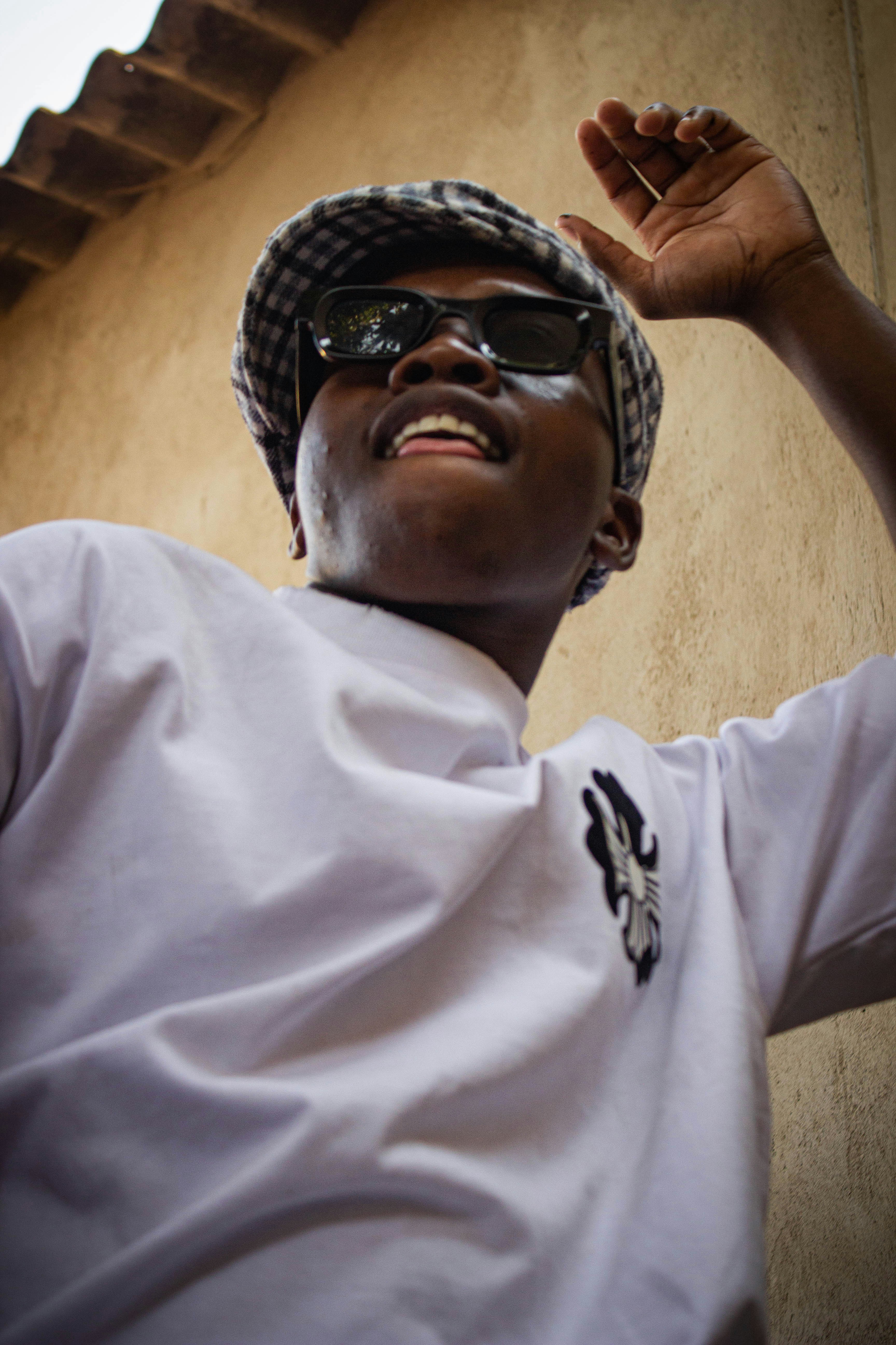 A person in a white t-shirt and plaid cap wearing sunglasses against a wall