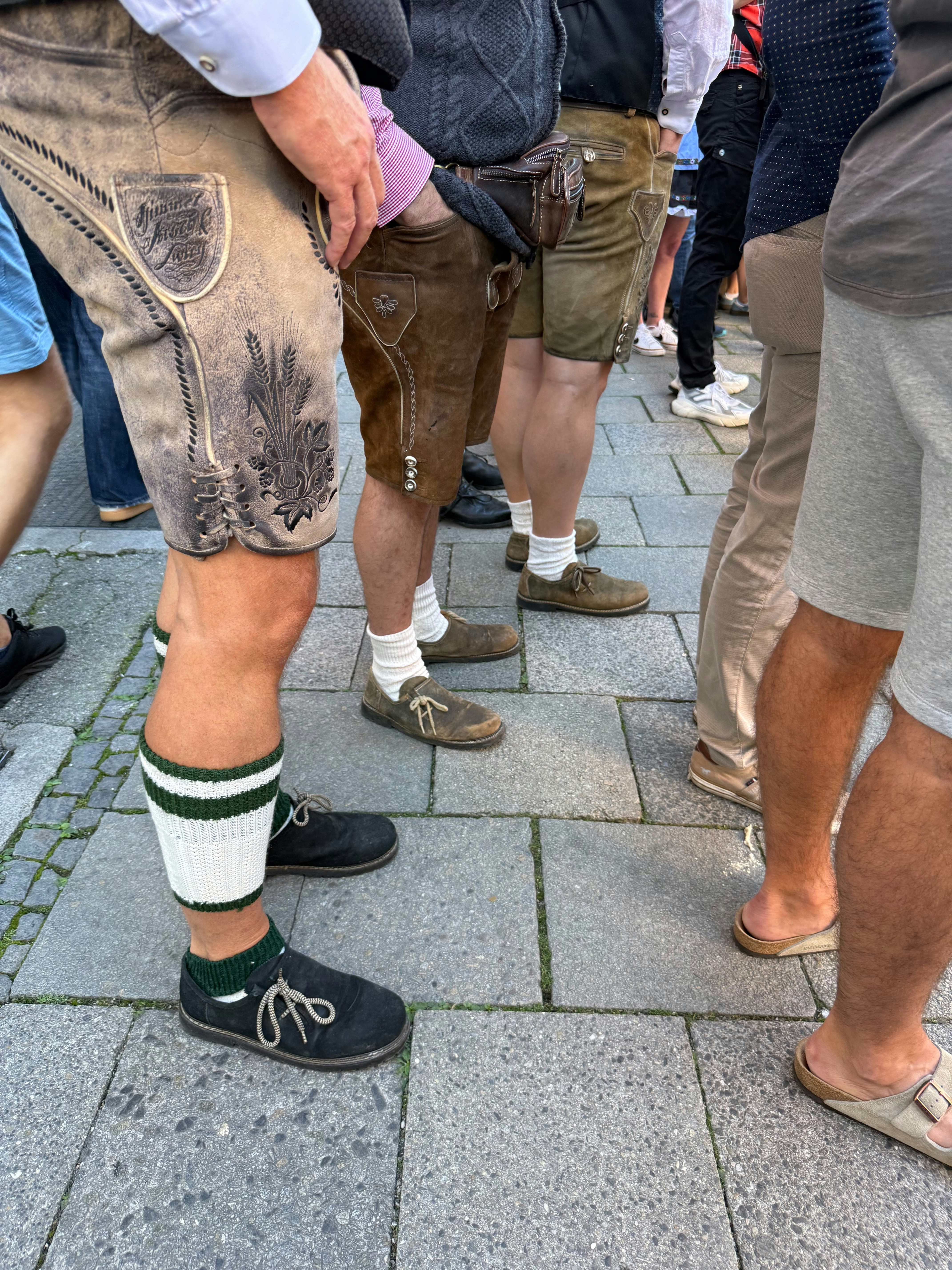 A group of people wearing traditional leather lederhosen standing on a stone paved street
