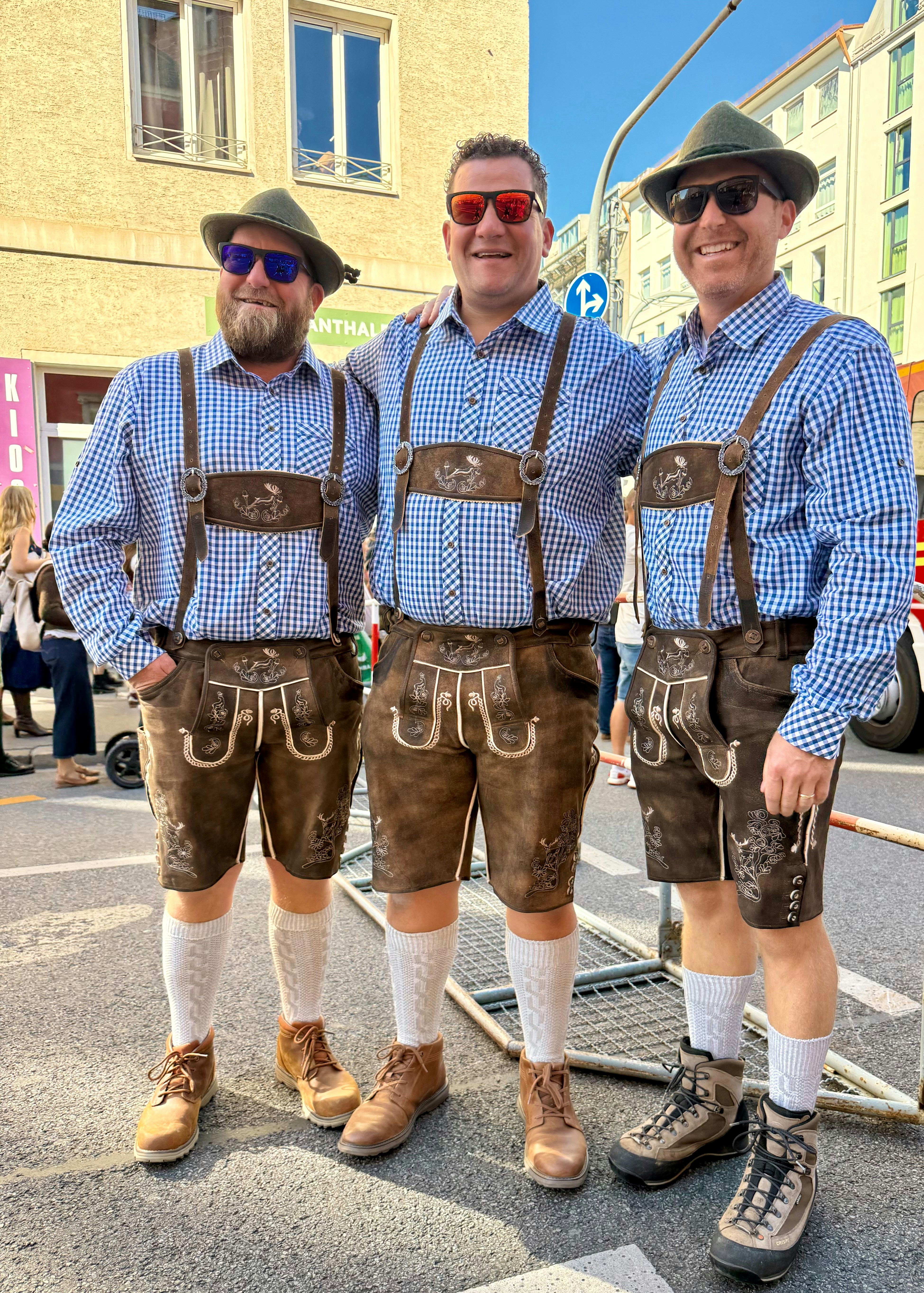 Three men in traditional lederhosen and blue checkered shirts standing on a city street