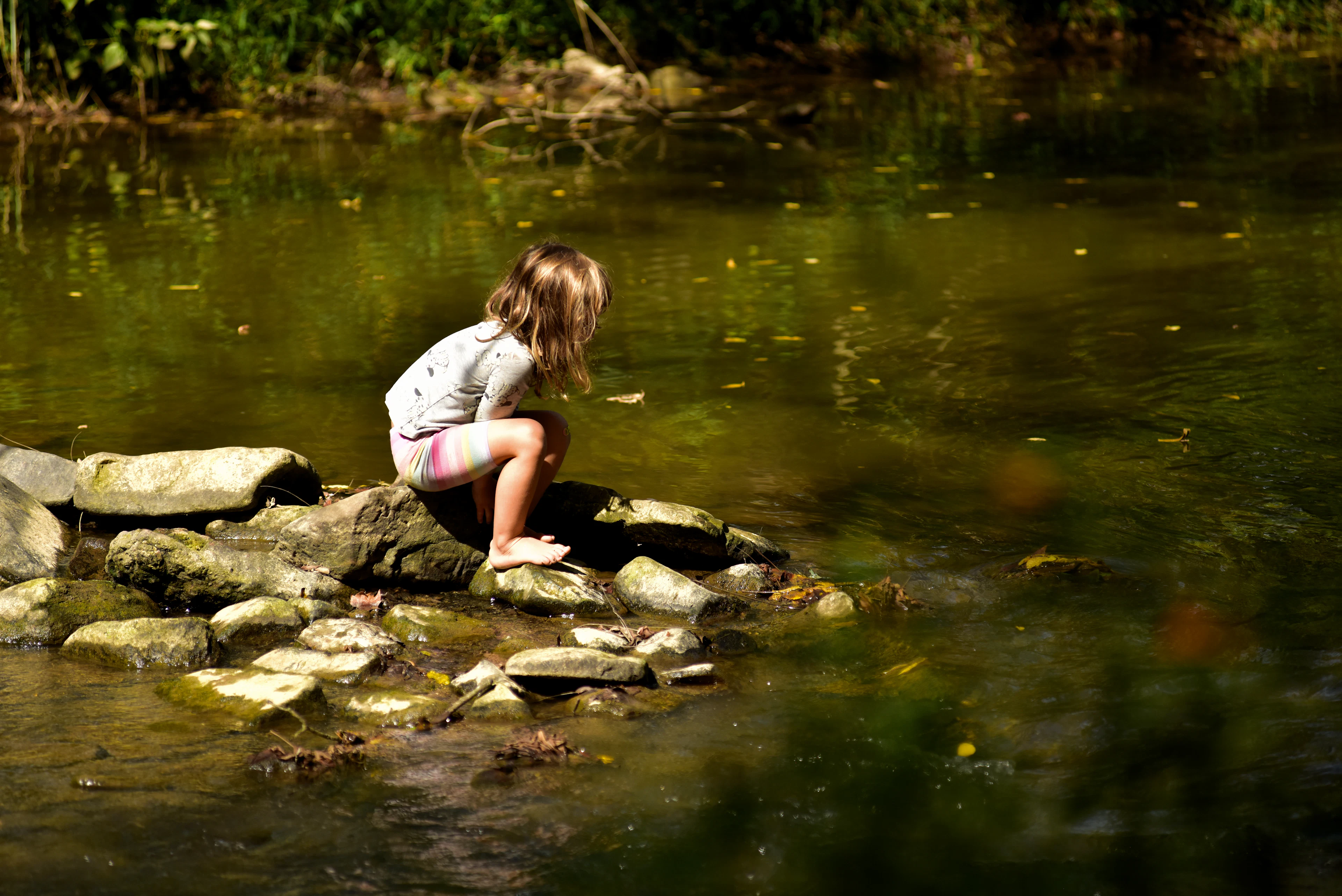 A child crouching on mossy rocks at the edge of a calm stream