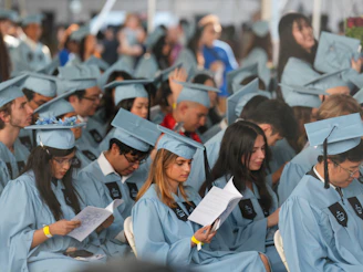 Graduates in light blue caps and gowns reading programs at a commencement ceremony