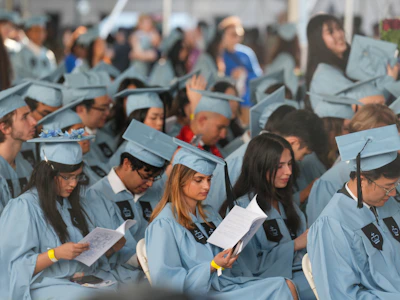 Graduates in light blue caps and gowns reading programs at a commencement ceremony