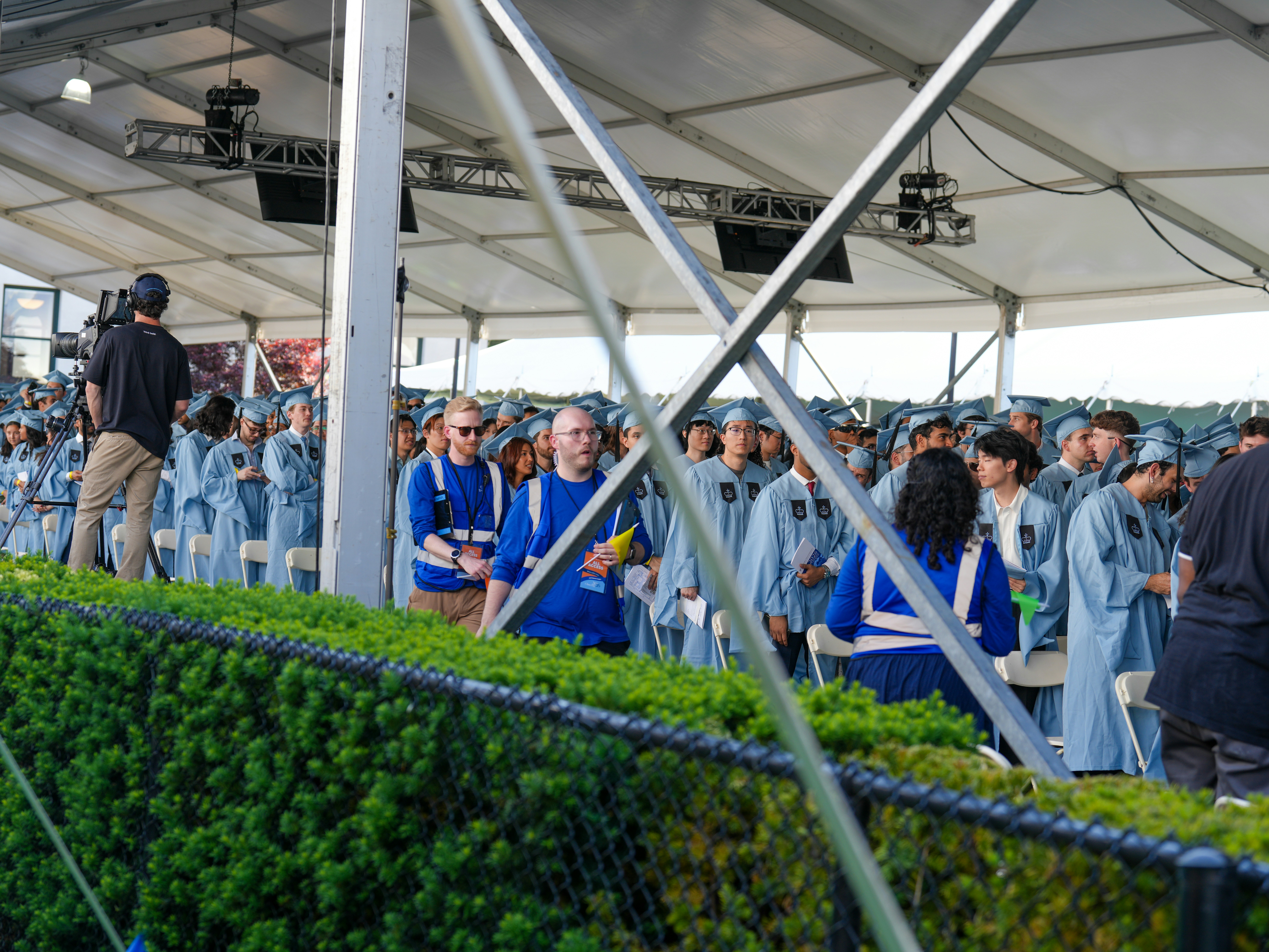 Seen through a chain-link fence and green hedge, rows of Columbia graduates in light blue gowns gather under a white ten