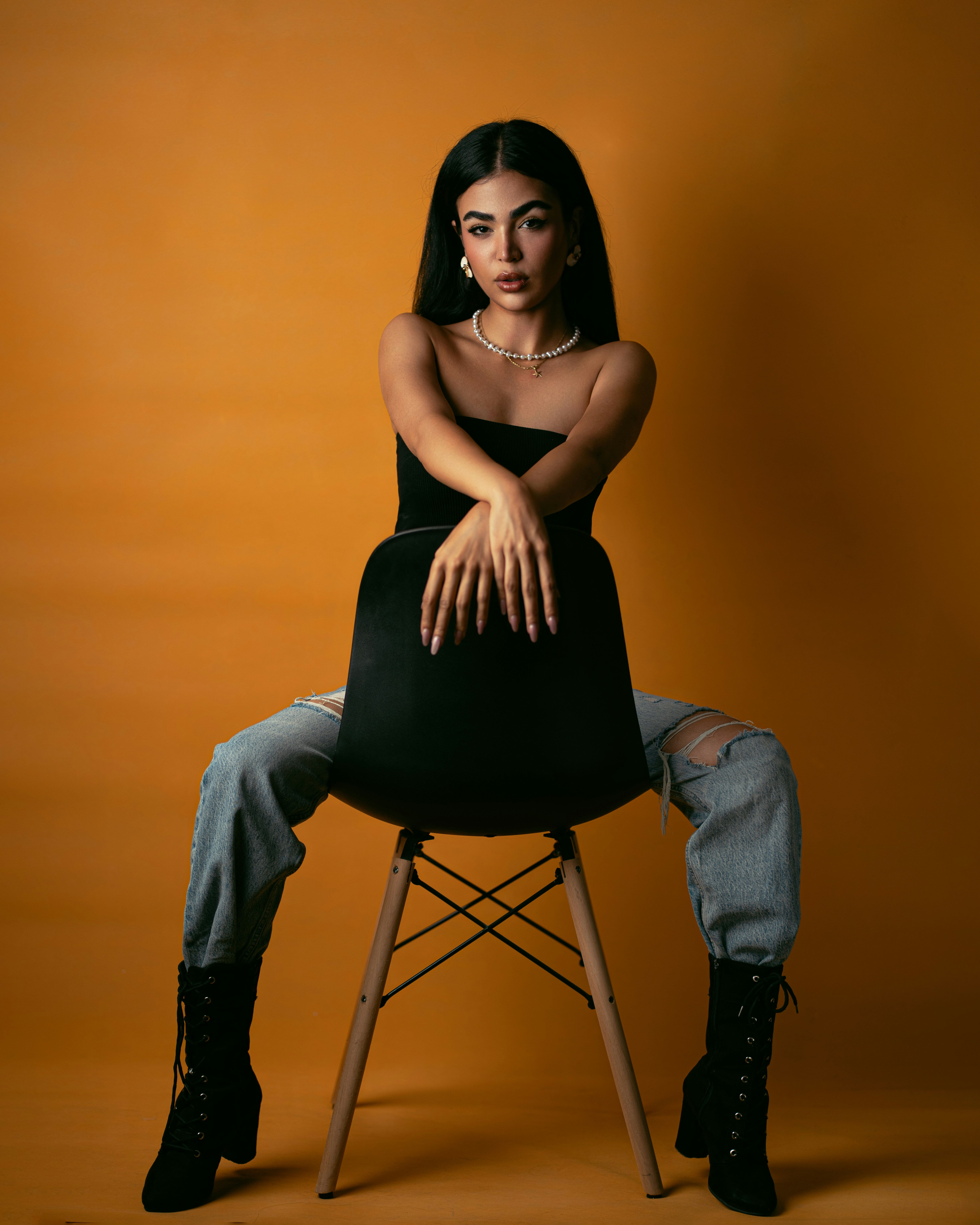 A woman in a black top and ripped jeans on a chair against orange