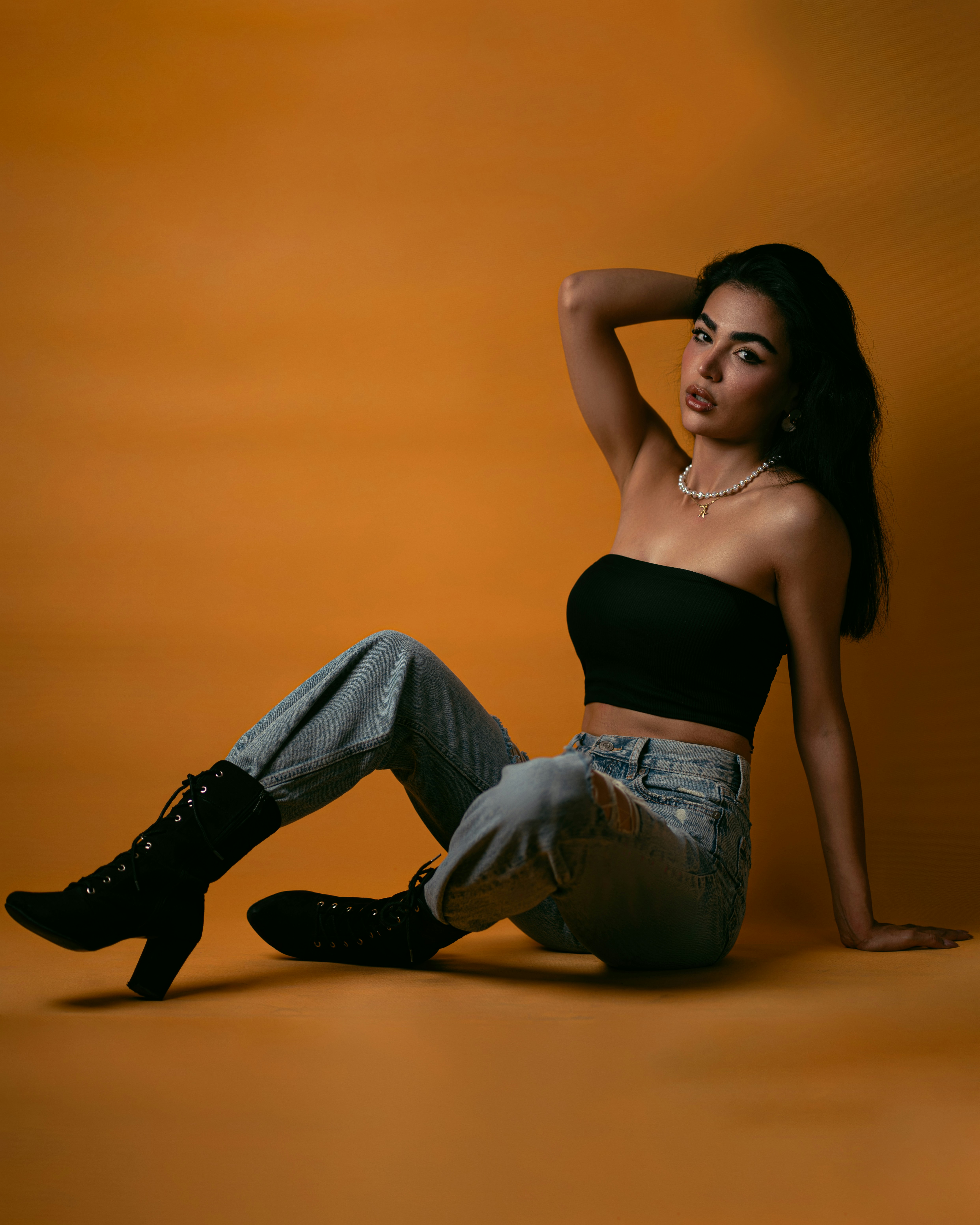 A woman in a black tube top and jeans sitting against an orange background