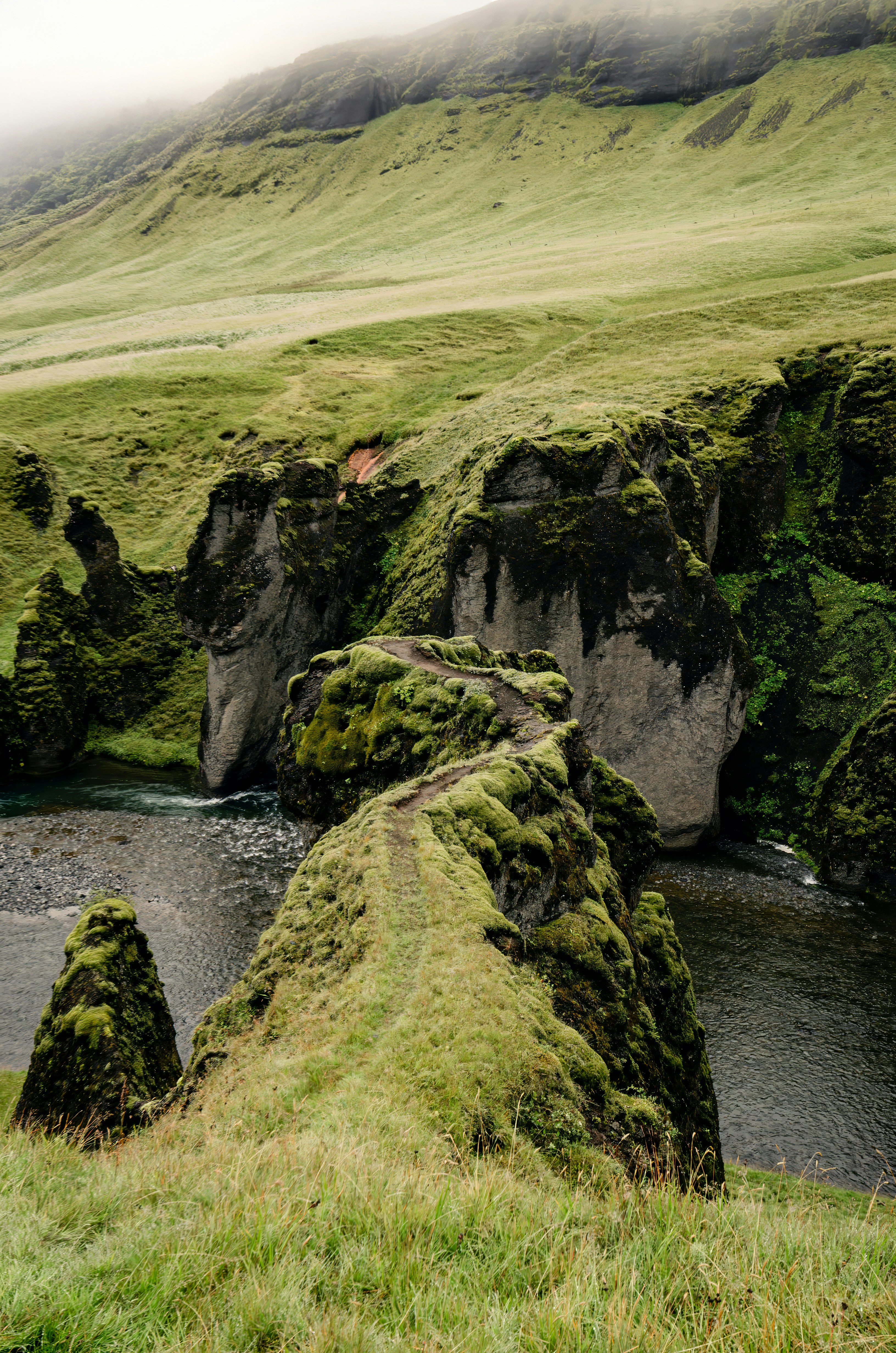 A narrow dirt path along a mossy ridge above a river in a valley