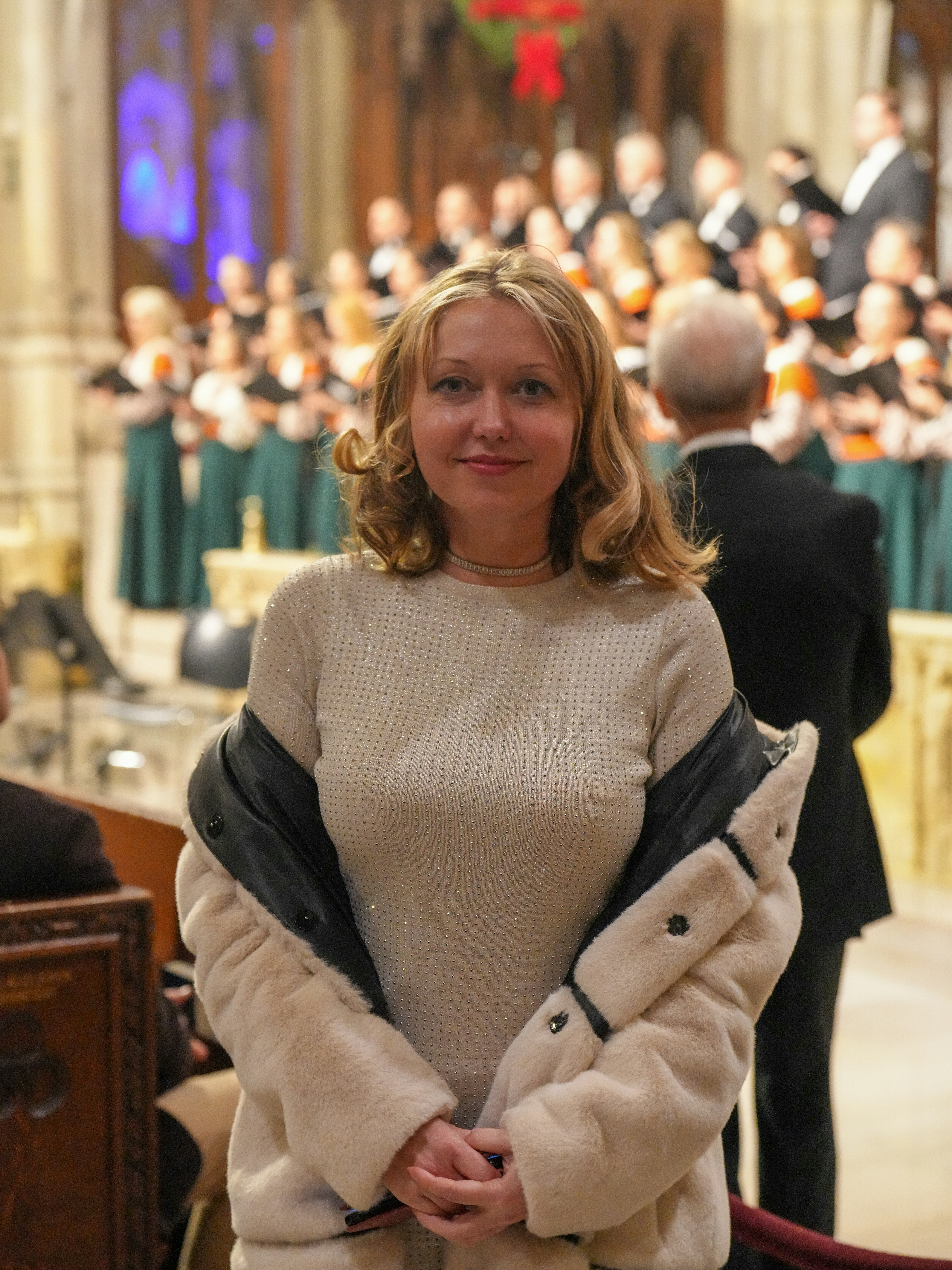 A woman in a beige sweater and faux fur coat before a choir
