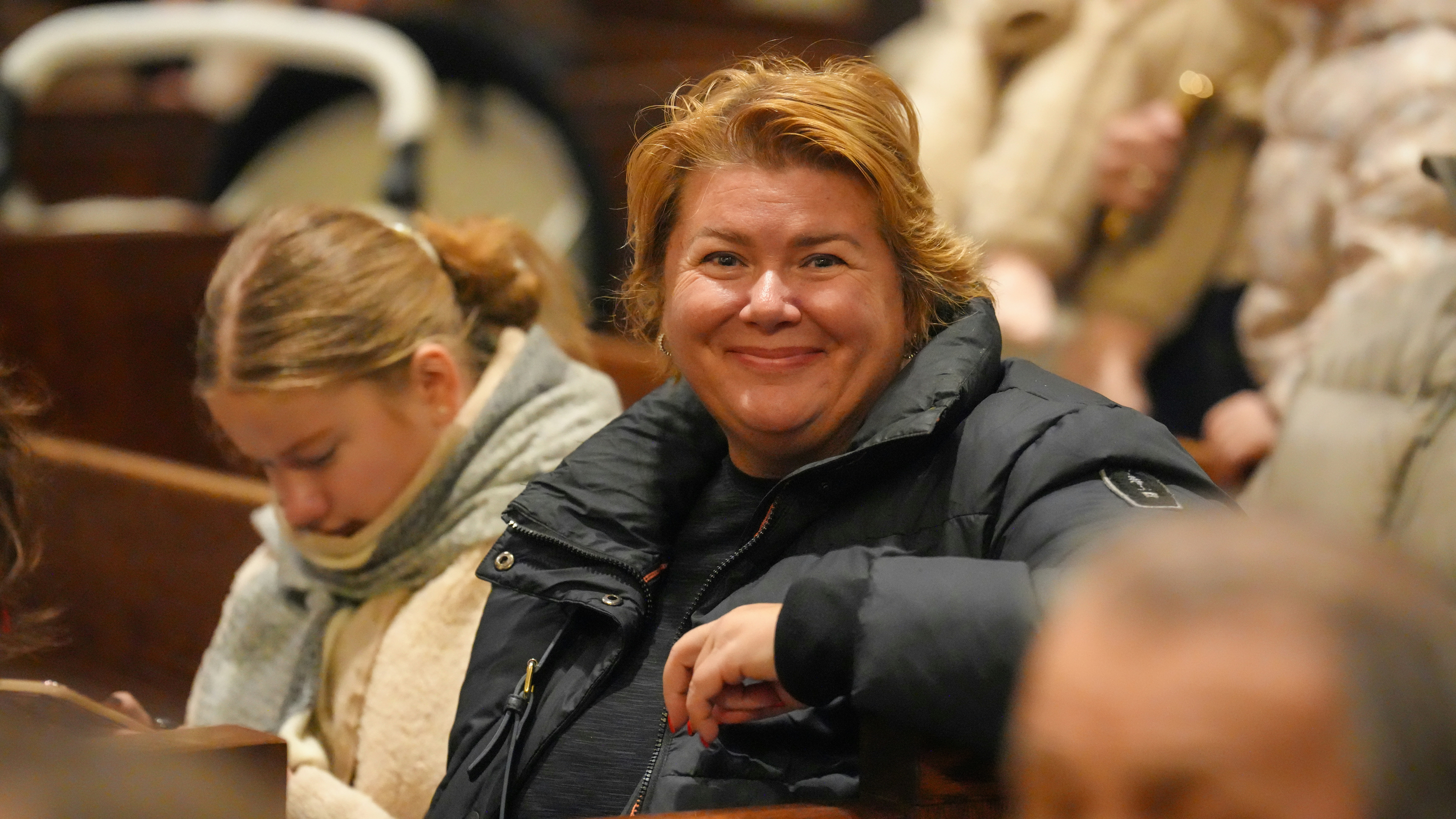 A smiling woman in a black puffer jacket sitting in a wooden pew