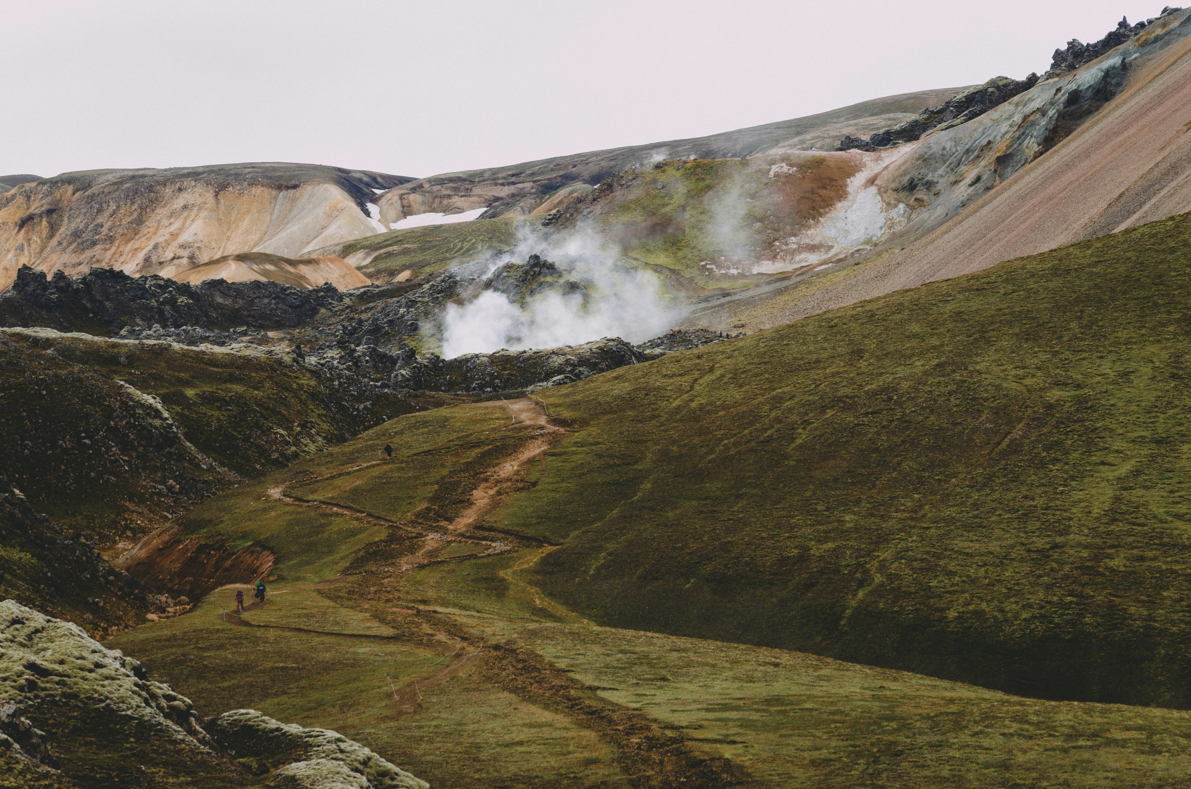 Hikers on a dirt path through green volcanic hills with rising geothermal steam