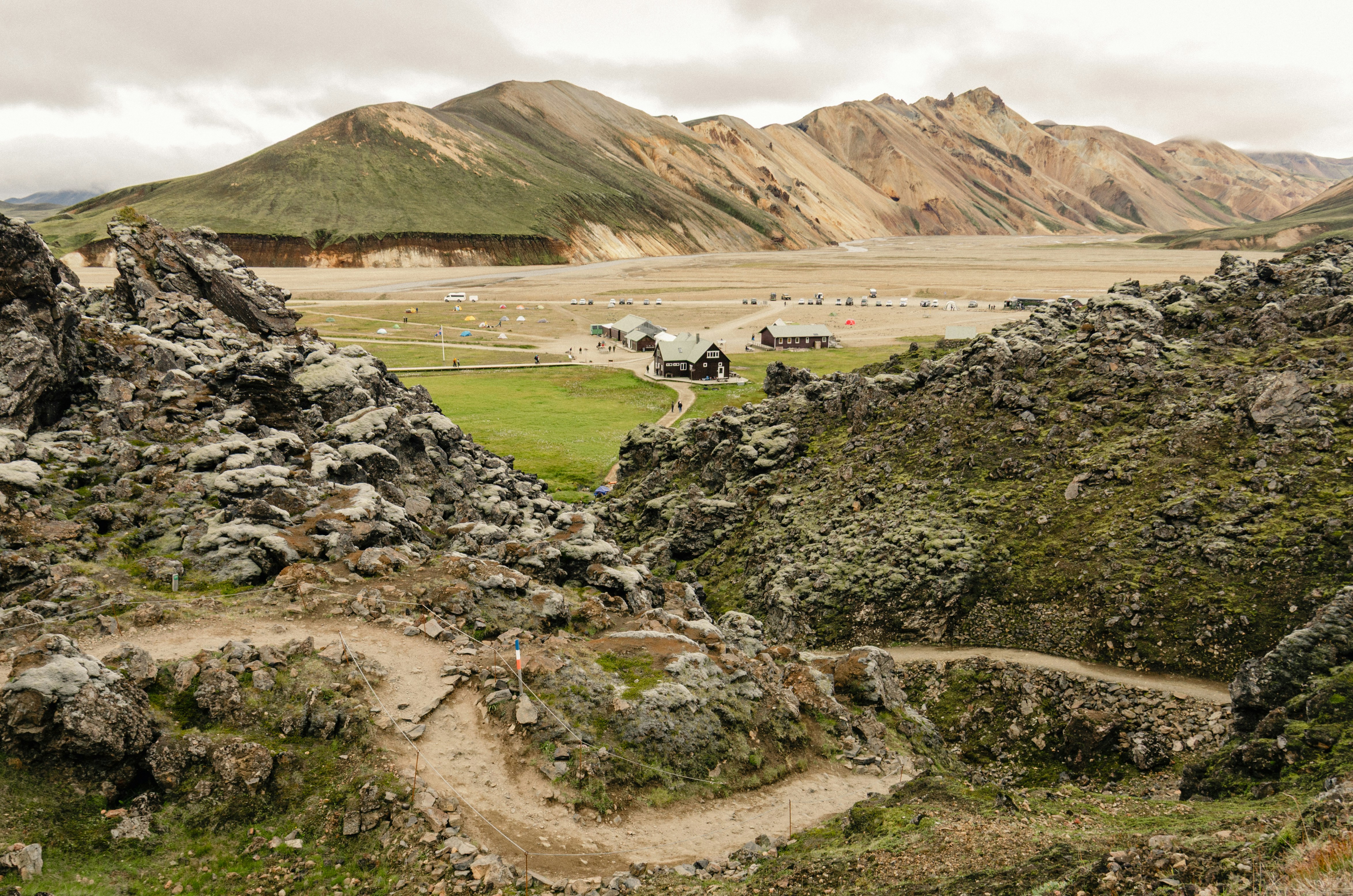 A winding trail through mossy volcanic rock leads down toward mountain huts and a campsite in a broad valley at Landmannalaugar, Iceland