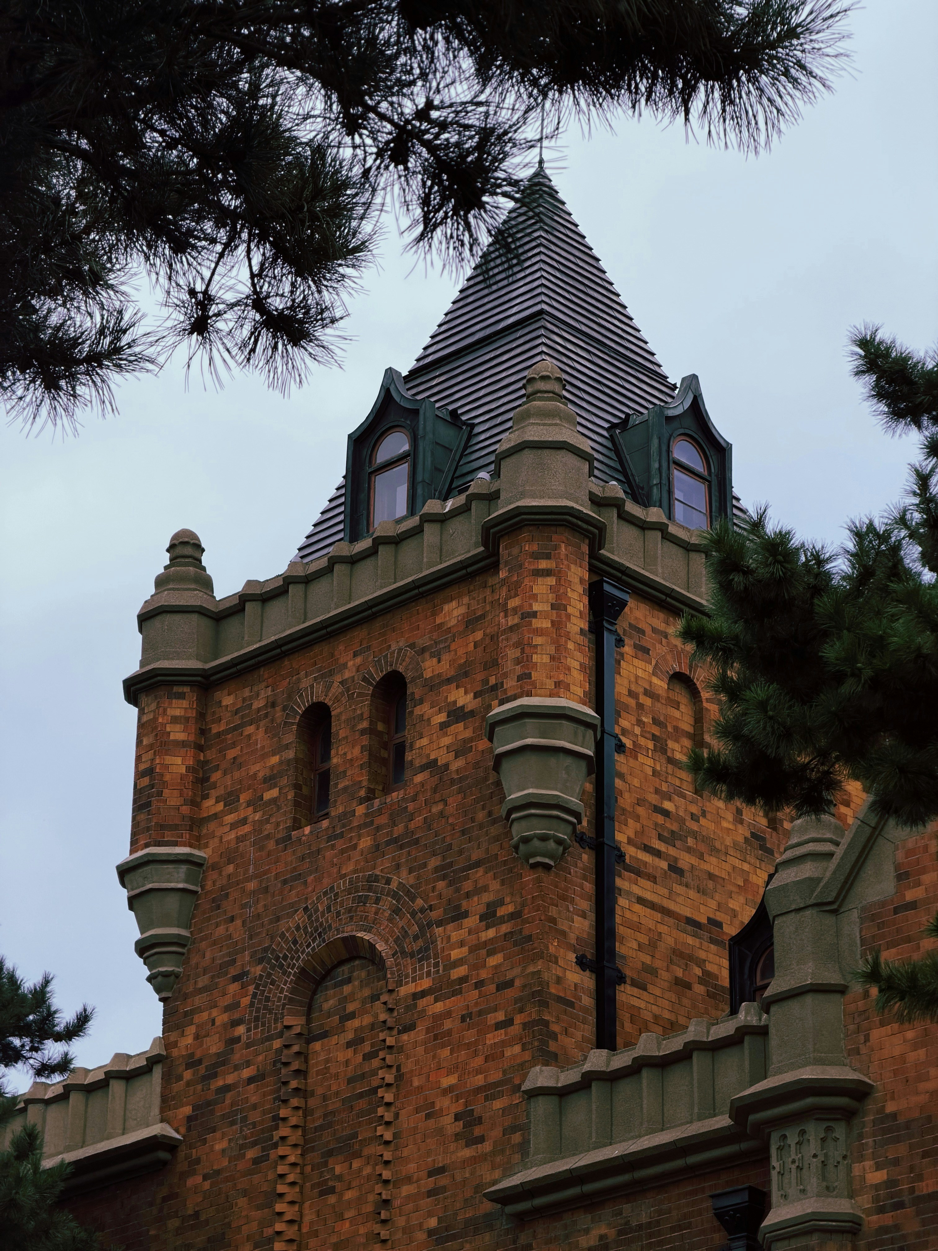 A brick tower with a dark roof and arched windows framed by pine branches