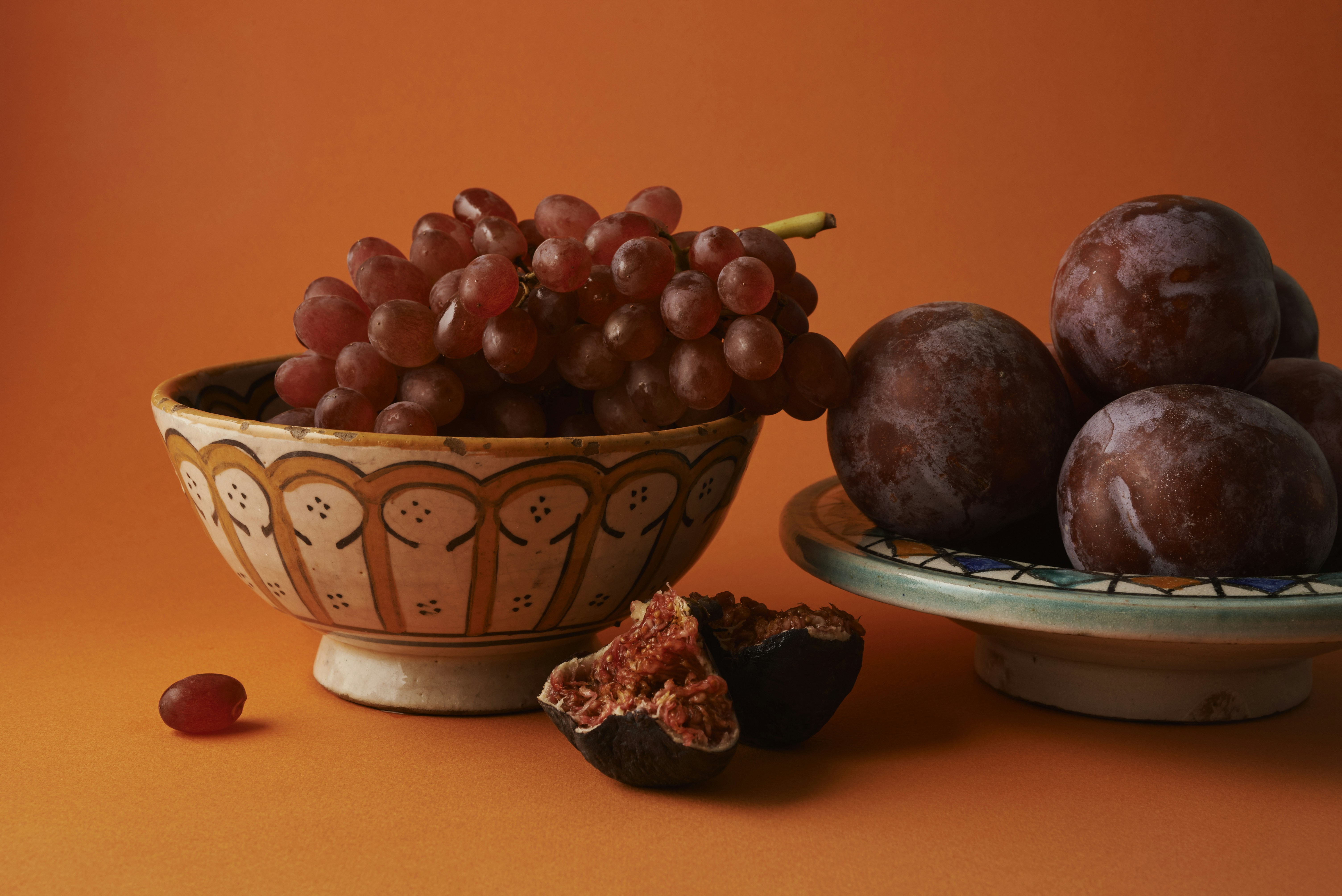 Red grapes, plums, and halved figs in ceramic bowls on an orange background