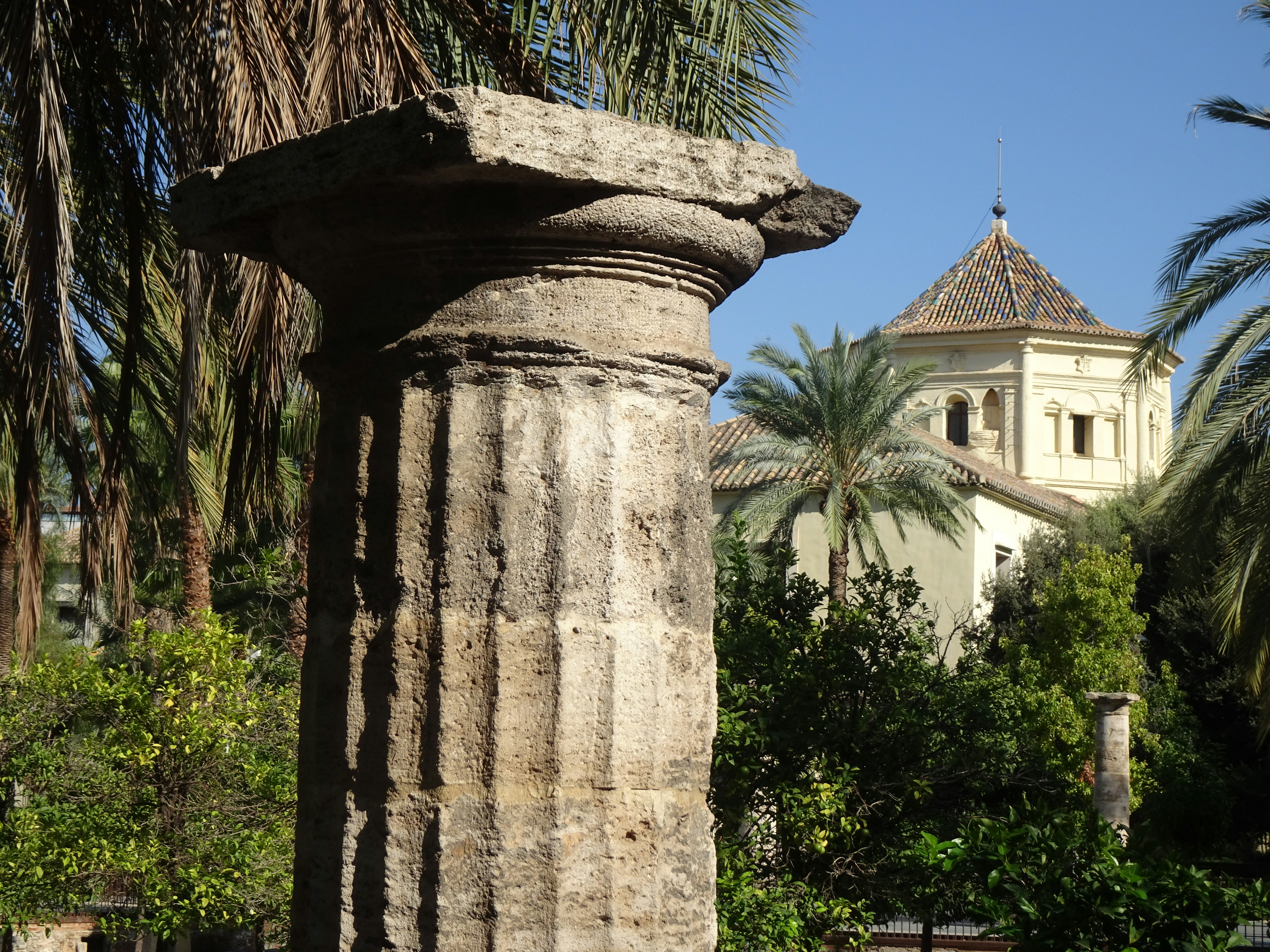A weathered stone column in a garden with palm trees and a domed building