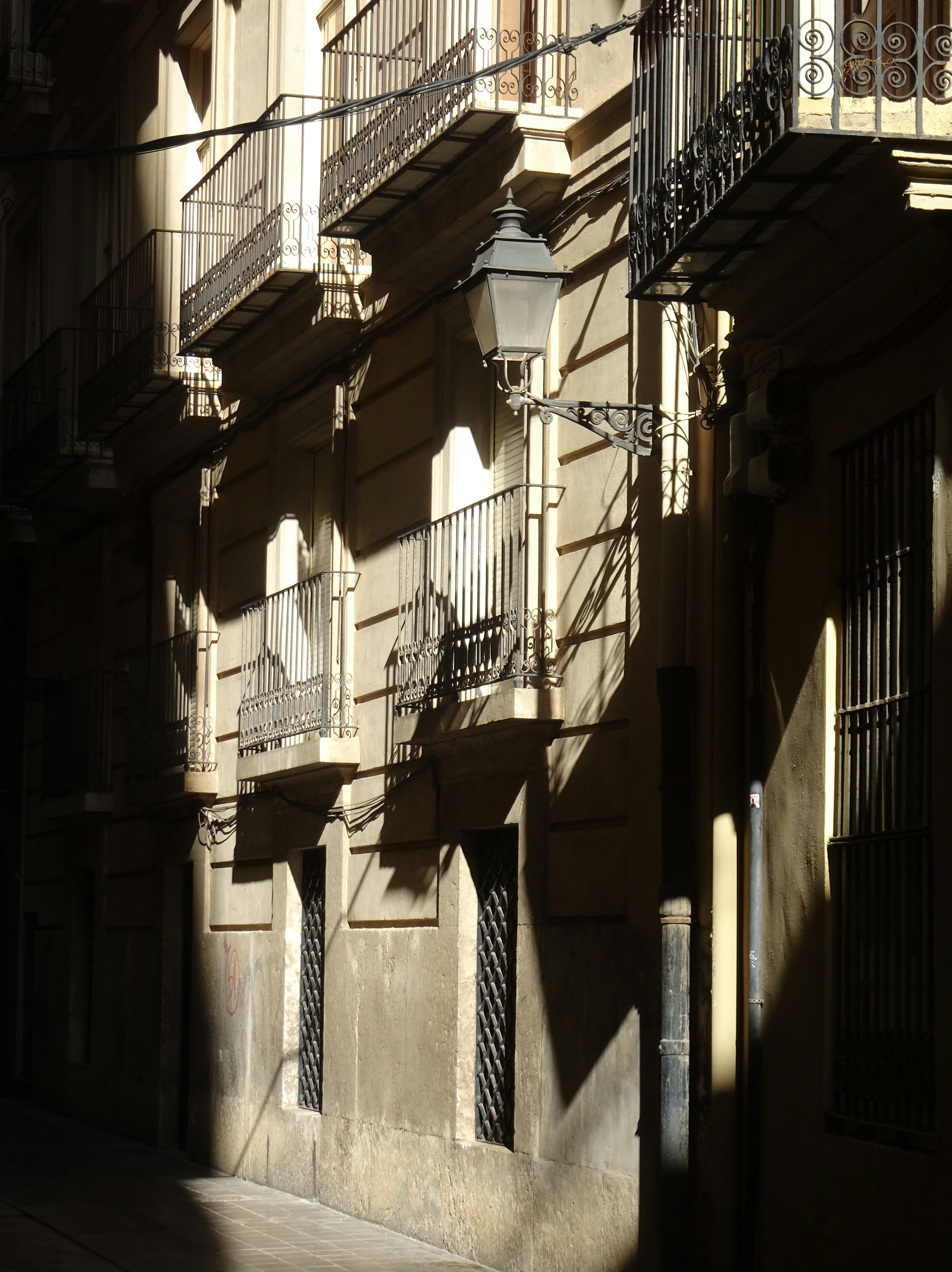A narrow street with beige buildings, iron balconies, and a wall-mounted lantern