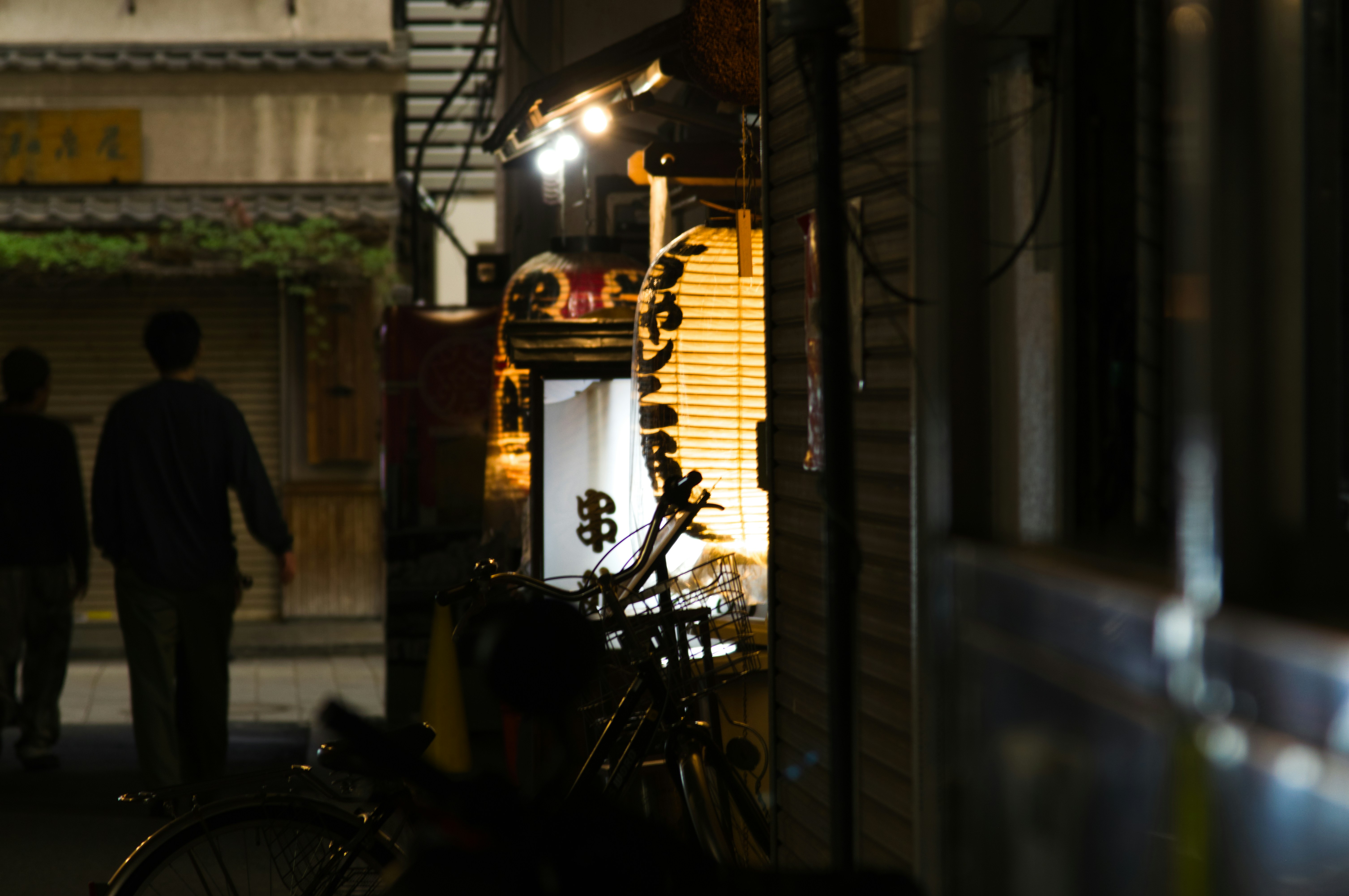 Two people walking down a narrow street in Japan past glowing paper lanterns