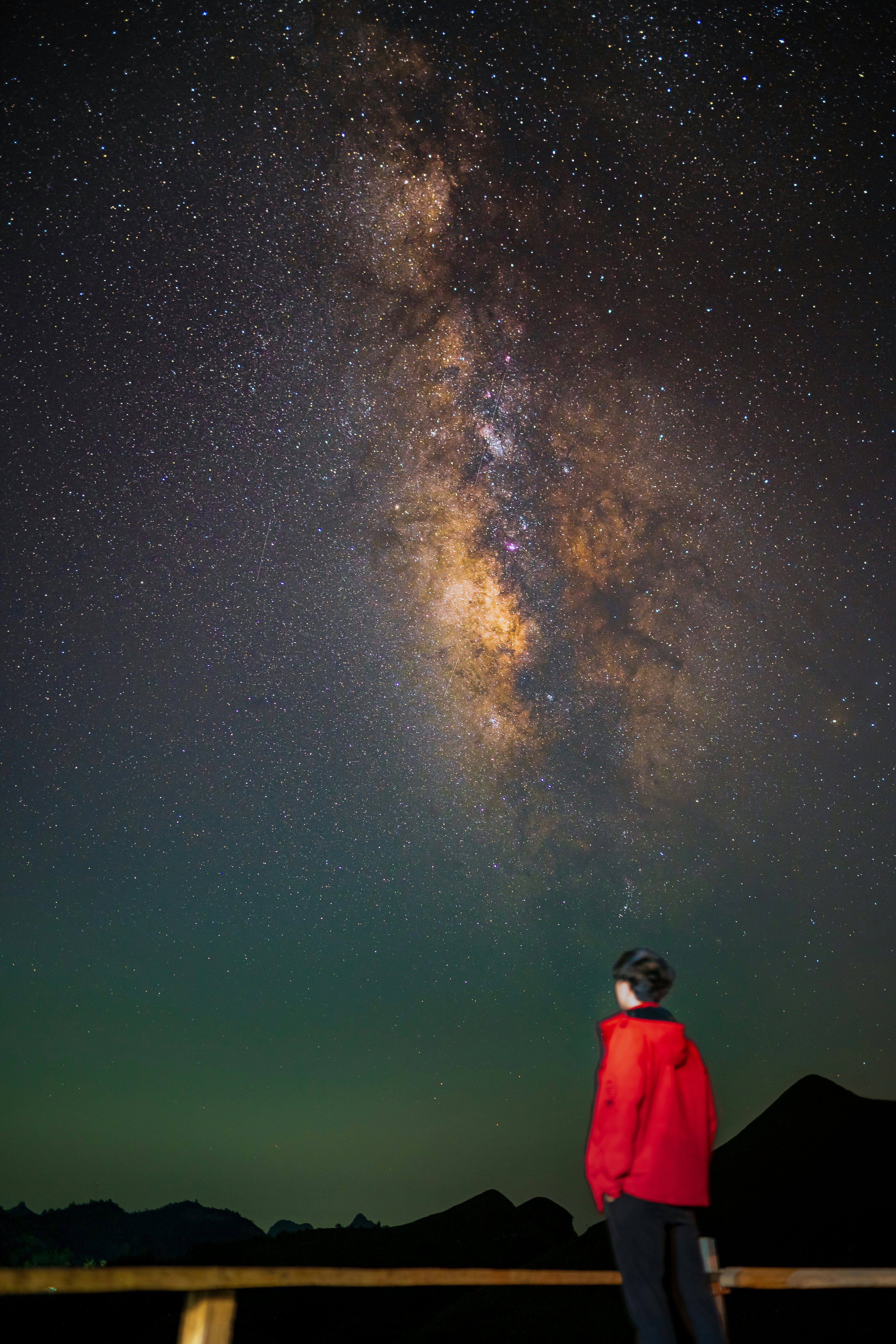A person in a red jacket looking at the Milky Way galaxy above mountains