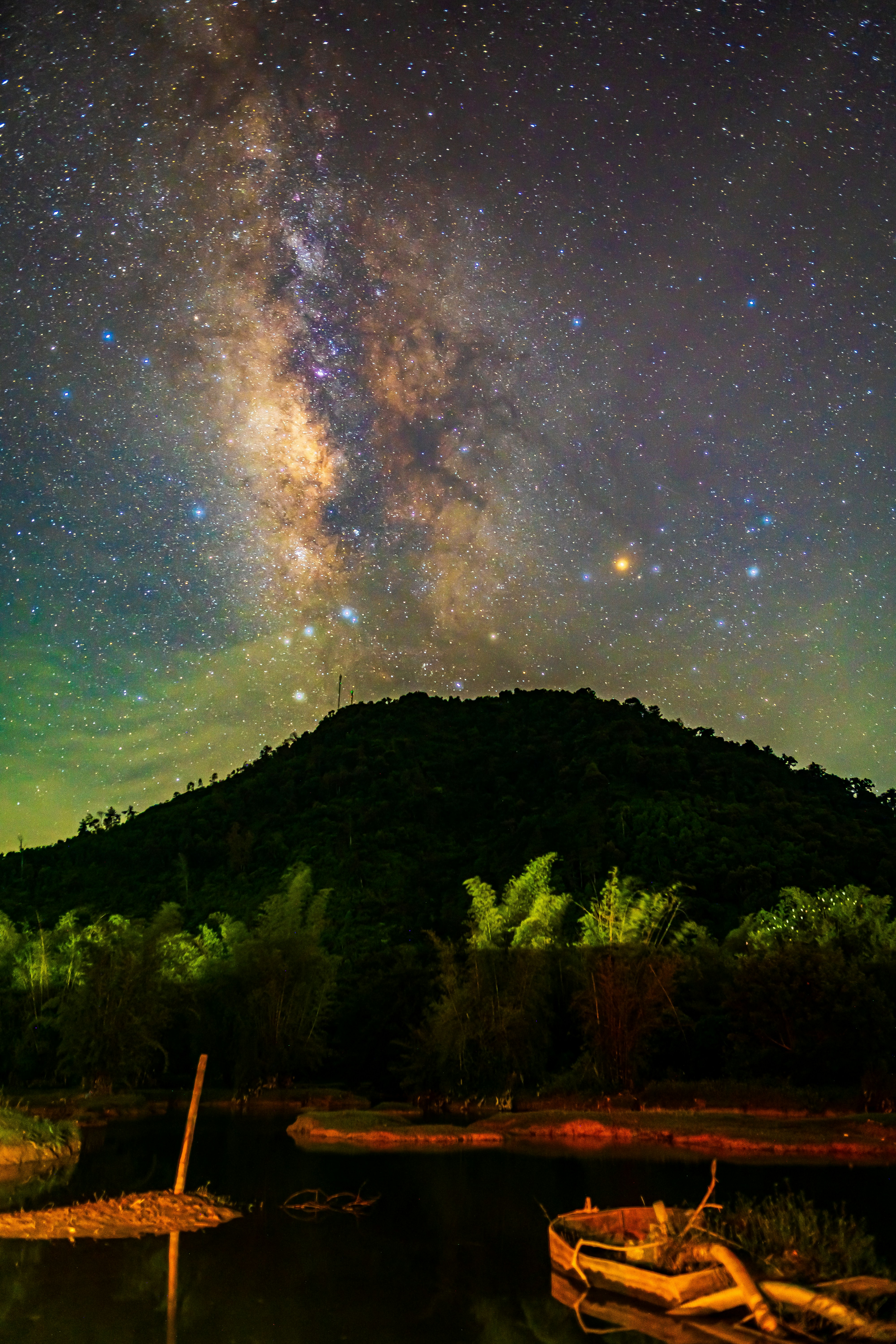 The Milky Way galaxy glowing above a dark forested hill and a small boat