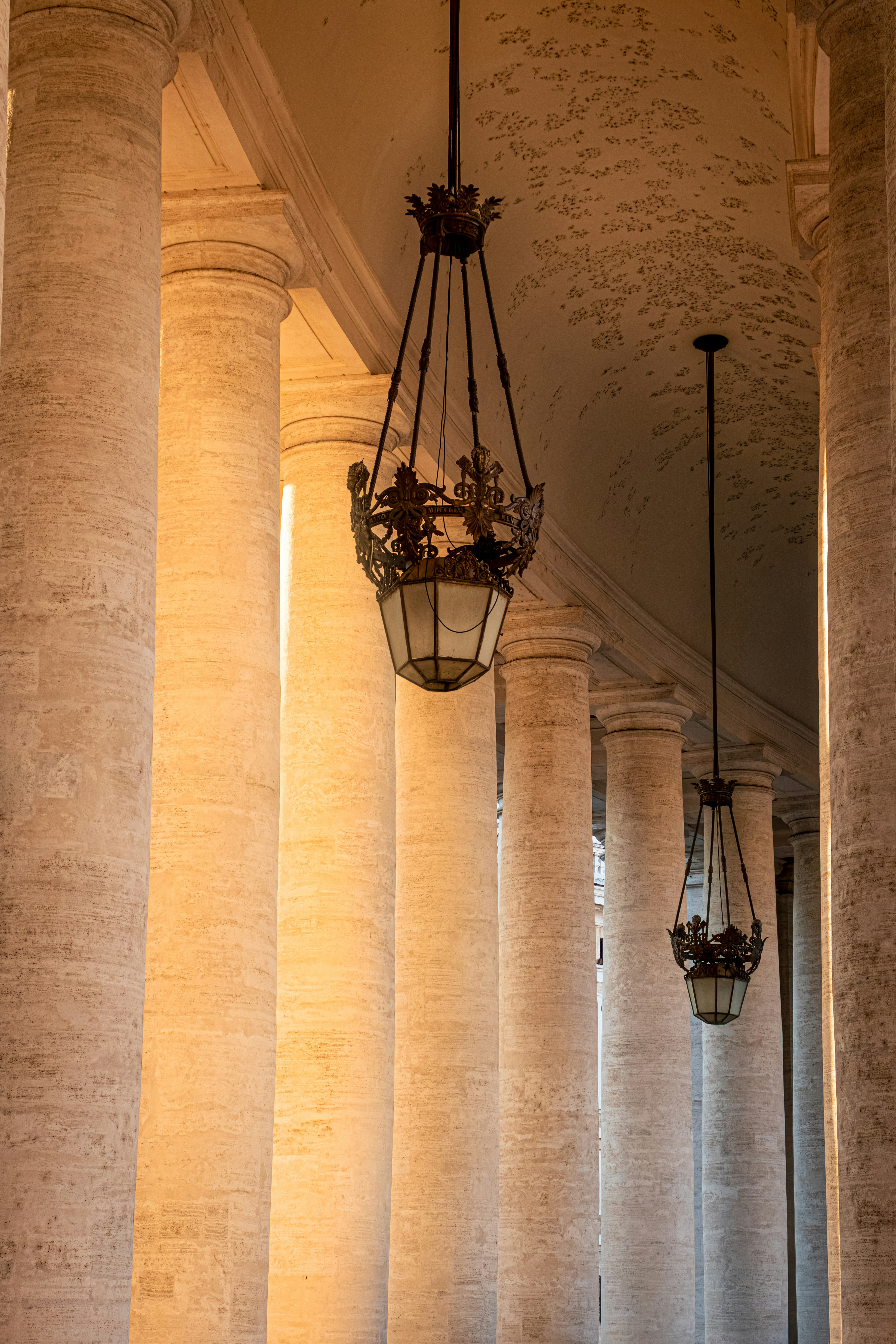 Ornate metal lanterns hanging between tall stone columns in a sunlit colonnade