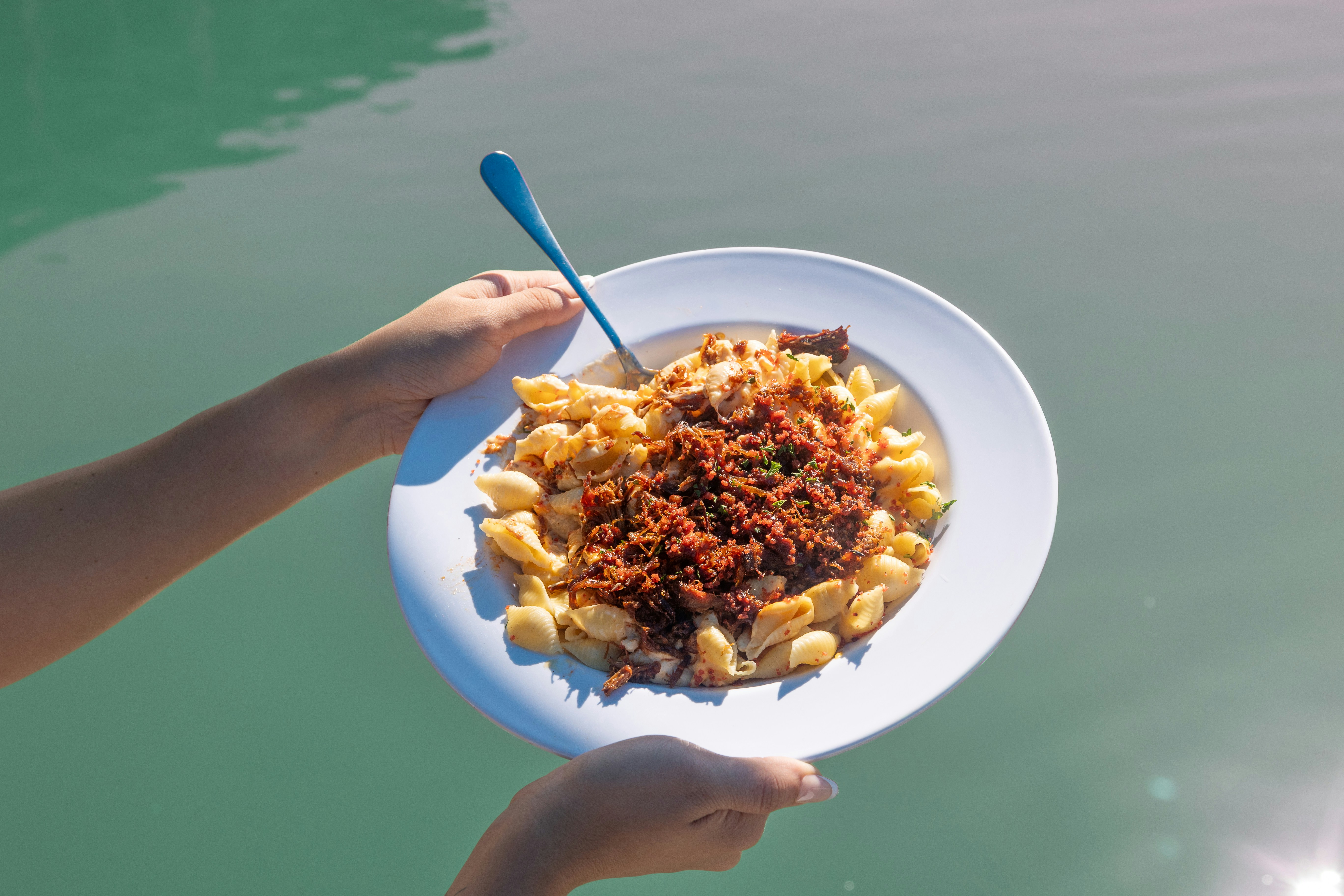 A vibrant horizontal perspective captures a diner holding a wide white plate of creamy pasta shells piled high with savory pulled barbecue meat and seasoning. Suspended directly above calm, sun-dappled turquoise water, the composition pairs indulgent comfort food with the refreshing ambiance of open-air waterfront dining.