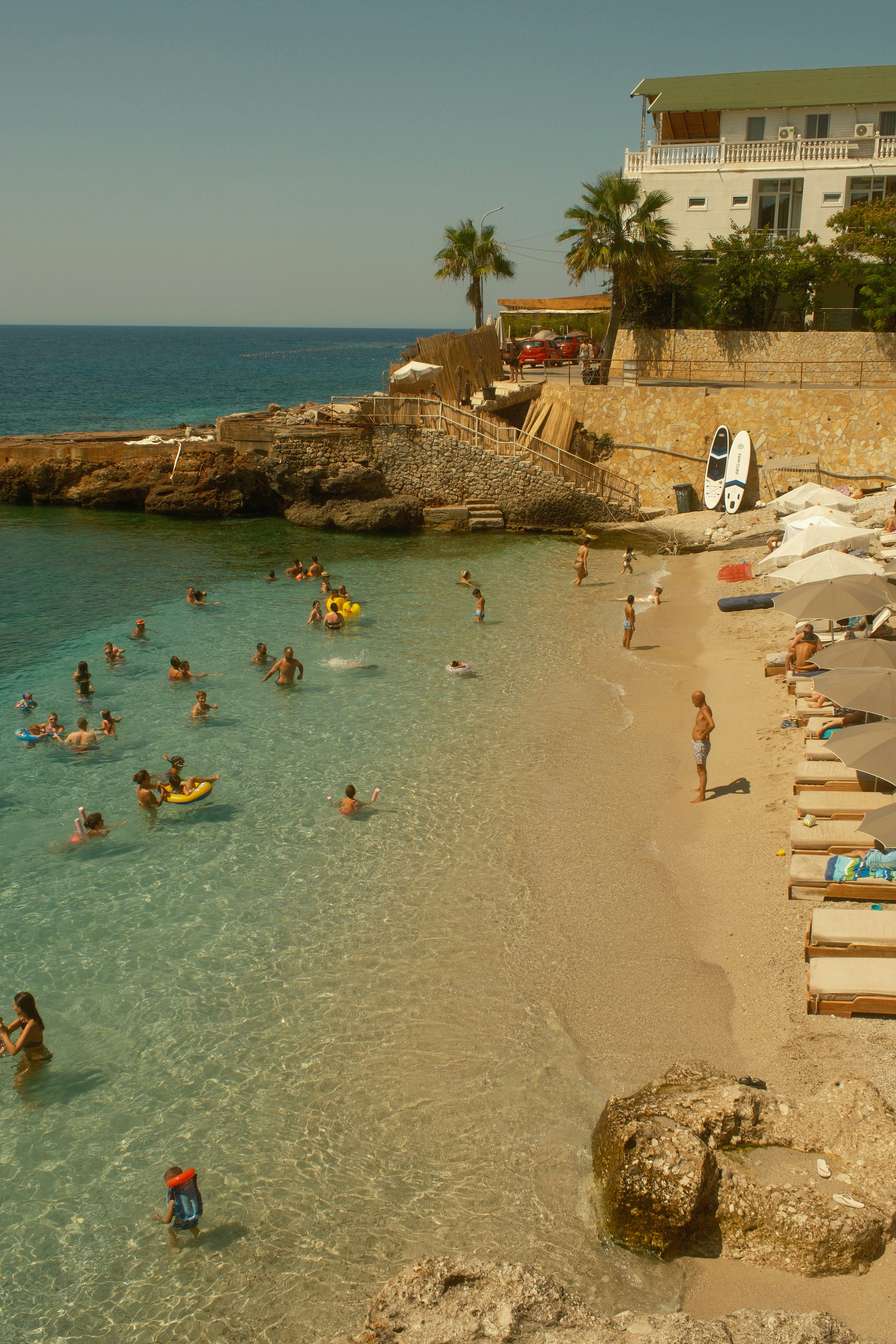 People swimming in clear turquoise water beside a sandy beach with lounge chairs