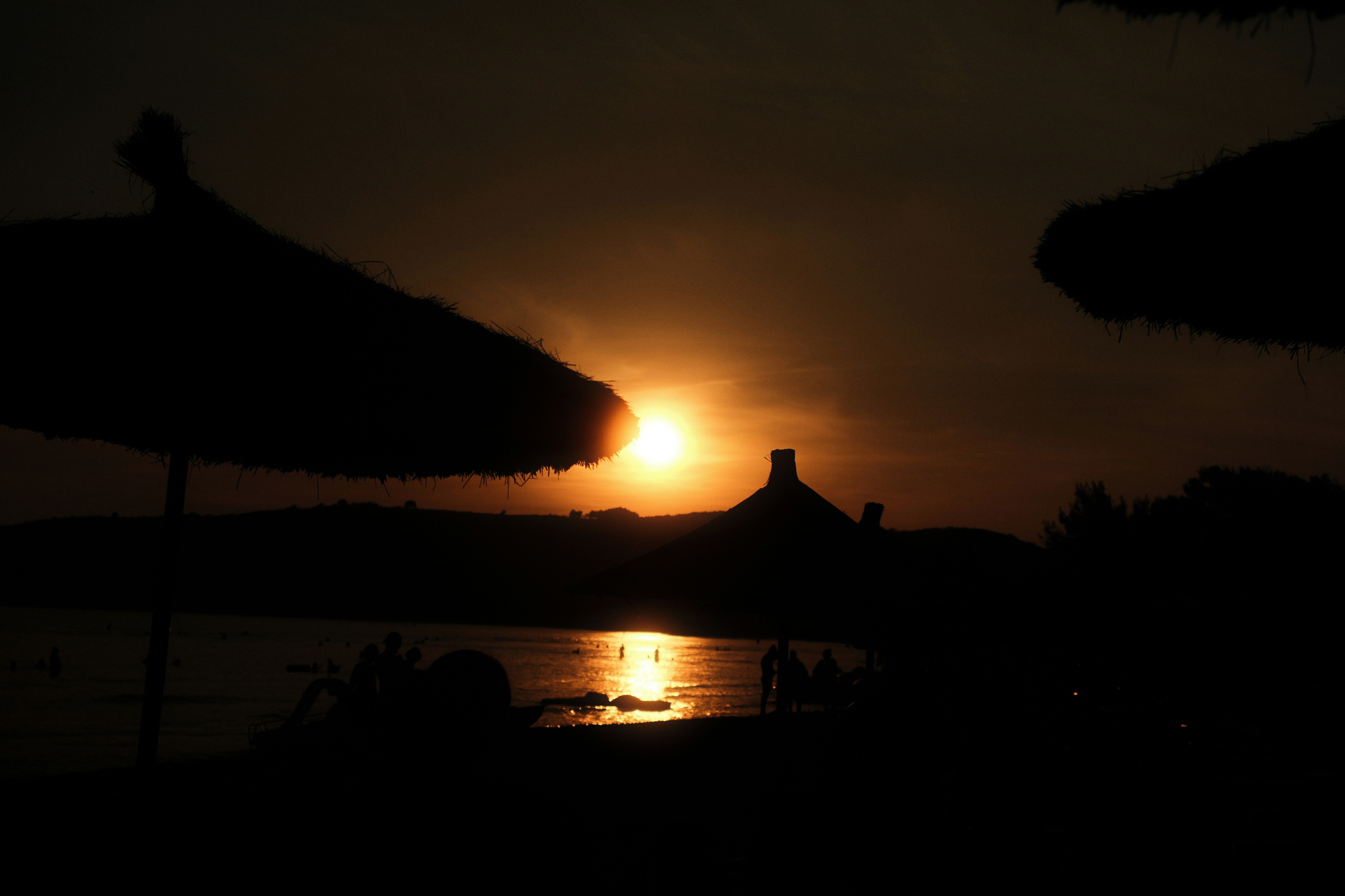 Silhouettes of straw umbrellas on a beach against a low sun and water