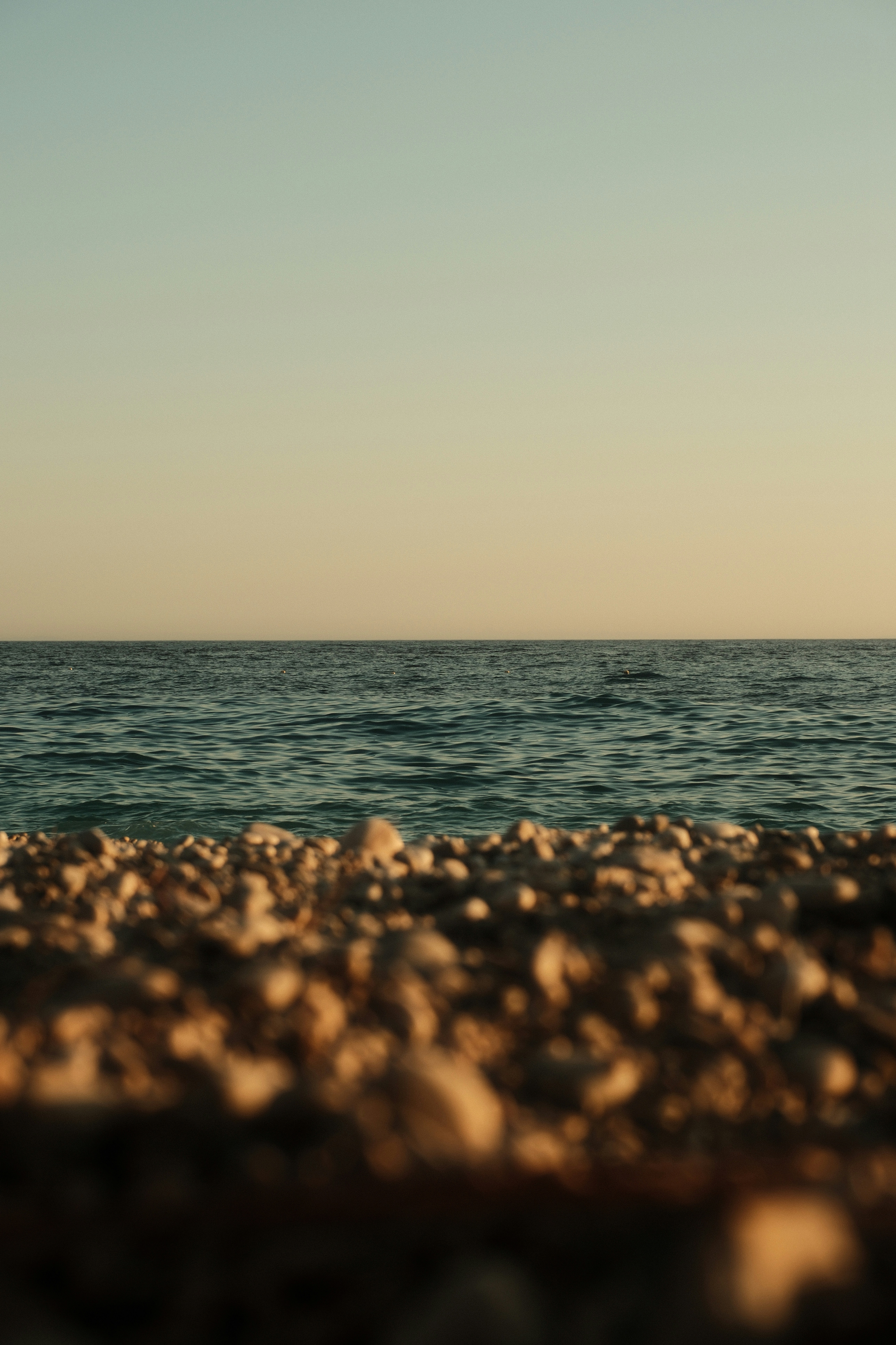 A pebble beach in the foreground with calm ocean water under a clear sky