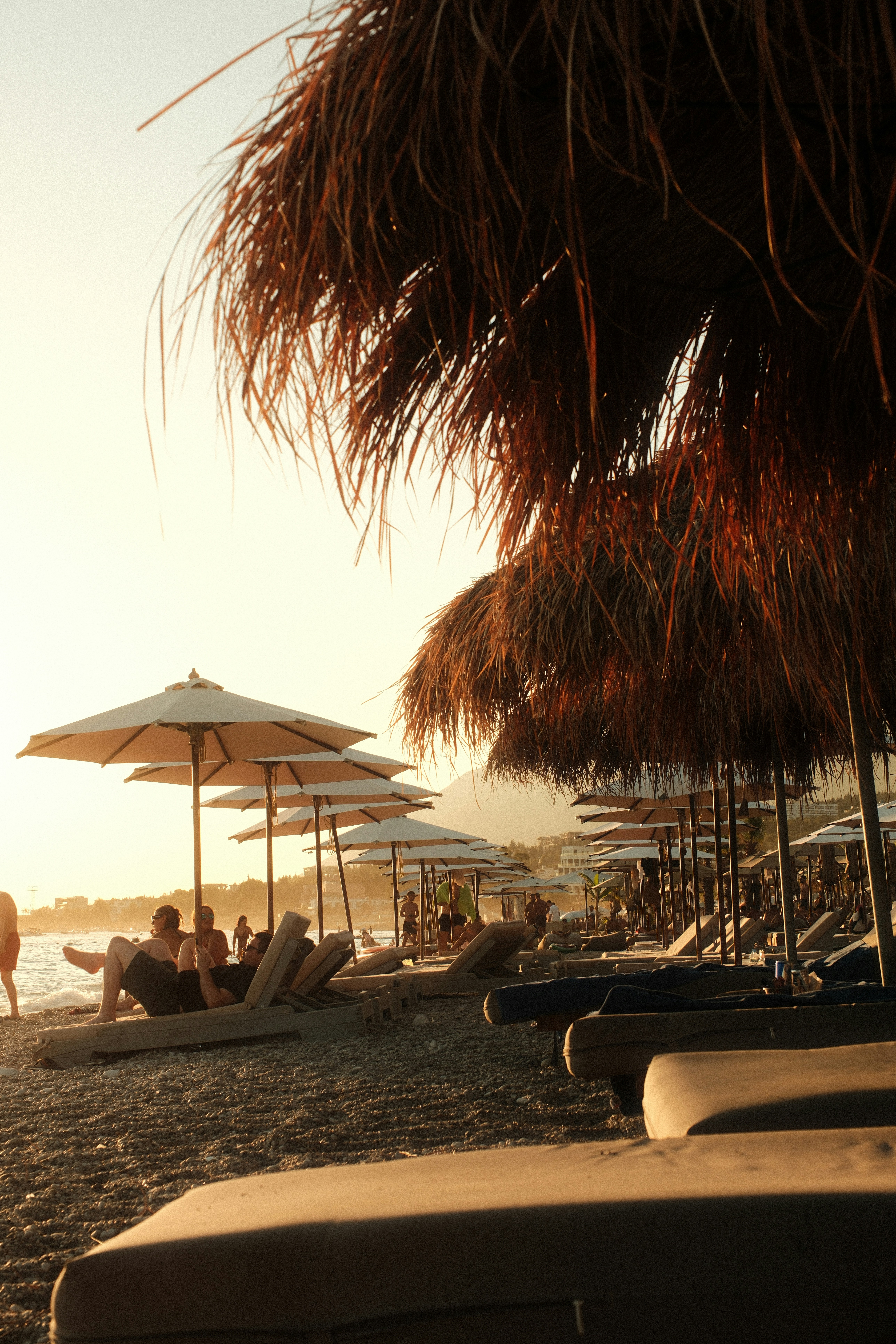 People relaxing on lounge chairs under straw umbrellas on a pebble beach at low sun