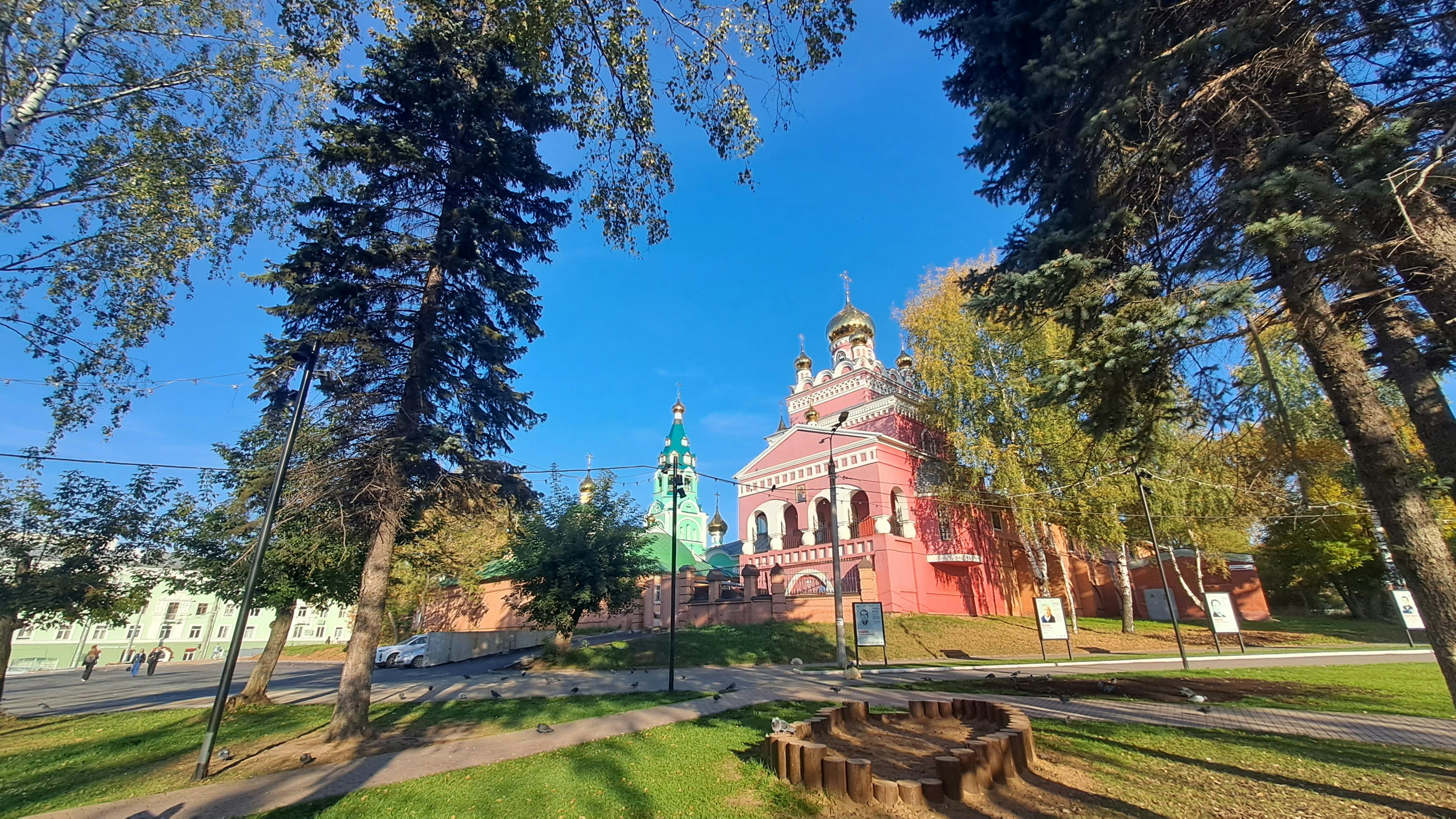 A pink Orthodox church with golden domes behind tall trees in a park