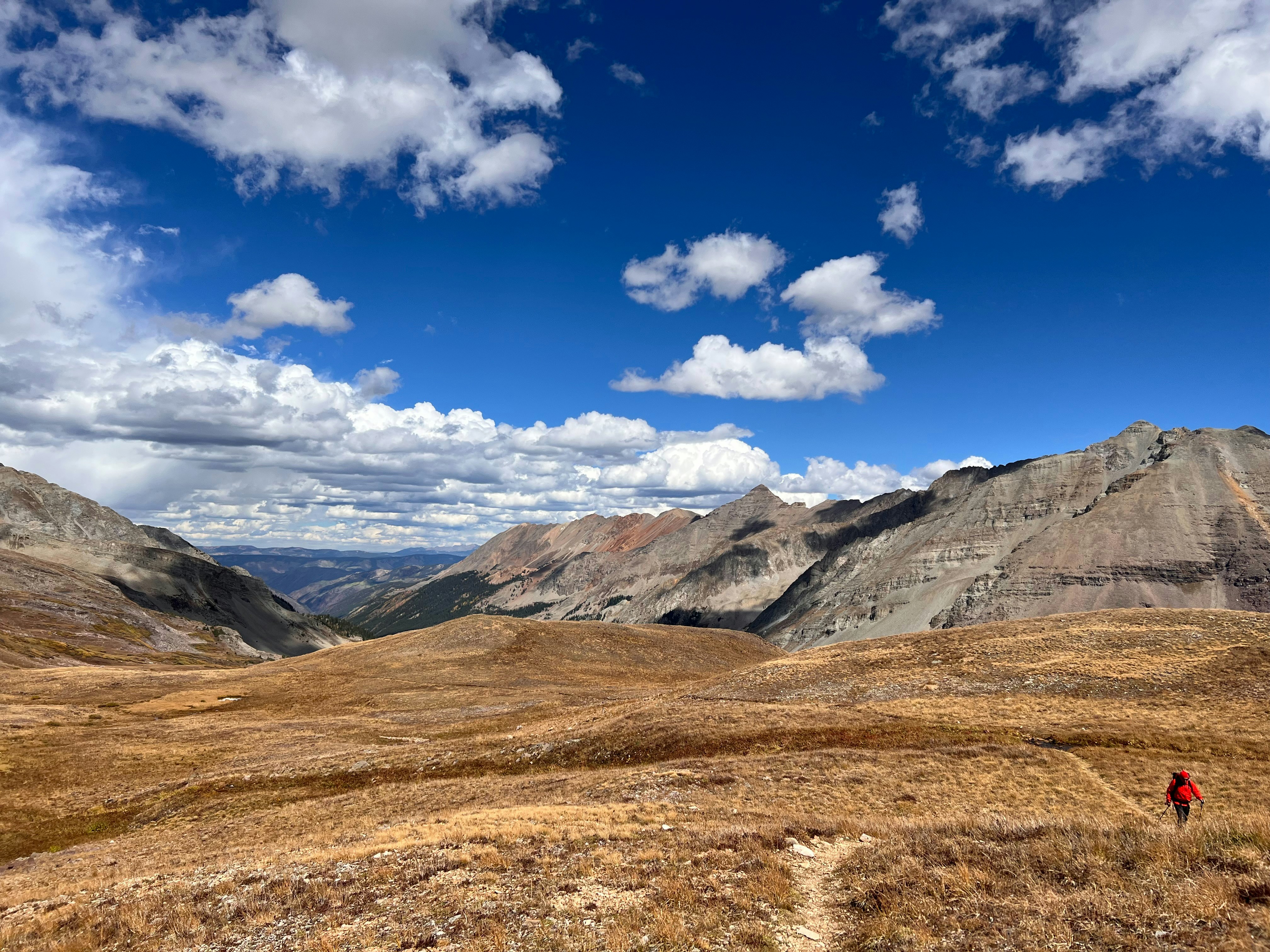 A hiker in a red jacket walking on a trail through a vast mountain landscape