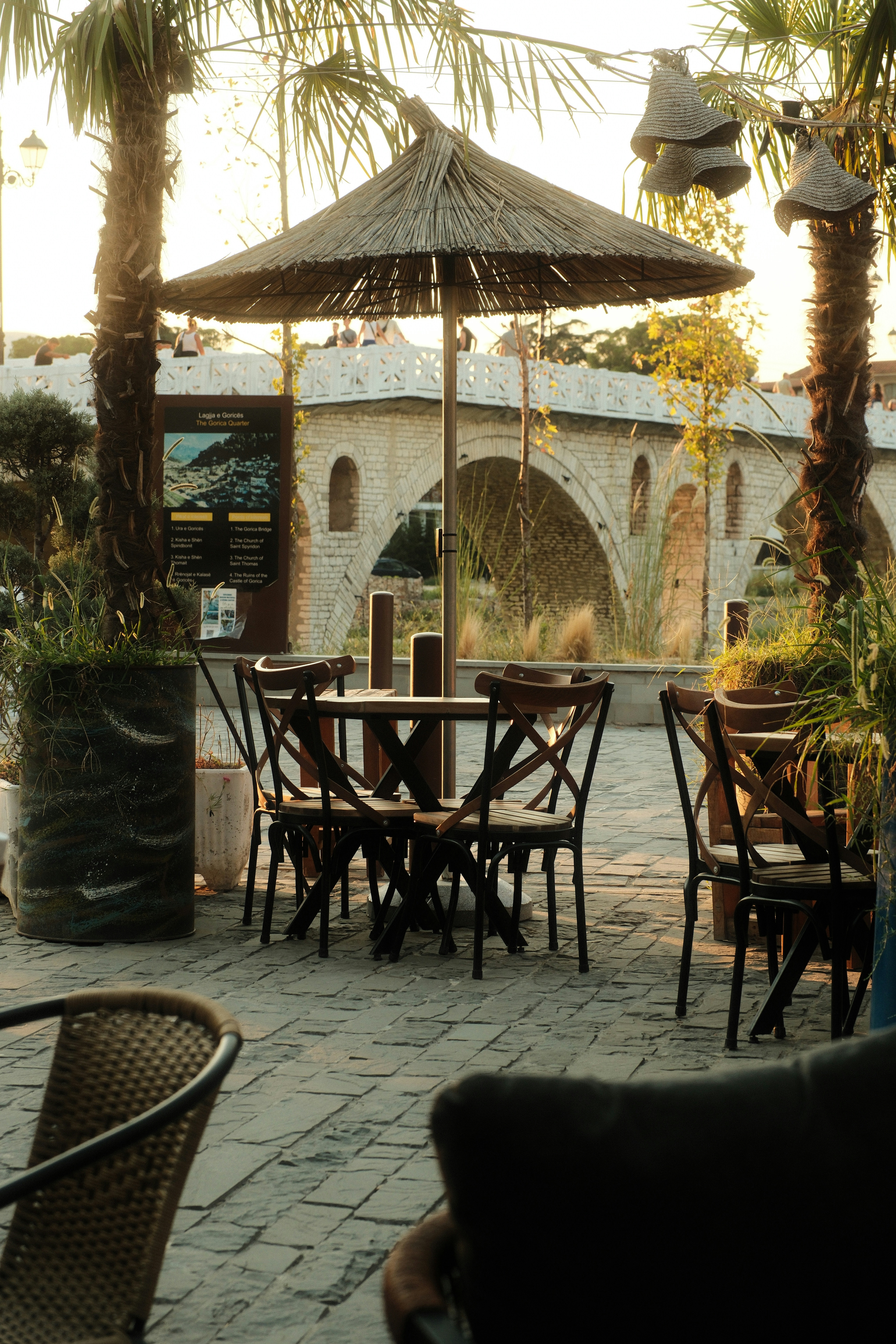 Outdoor cafe tables with straw umbrellas near a stone arch bridge in Berat