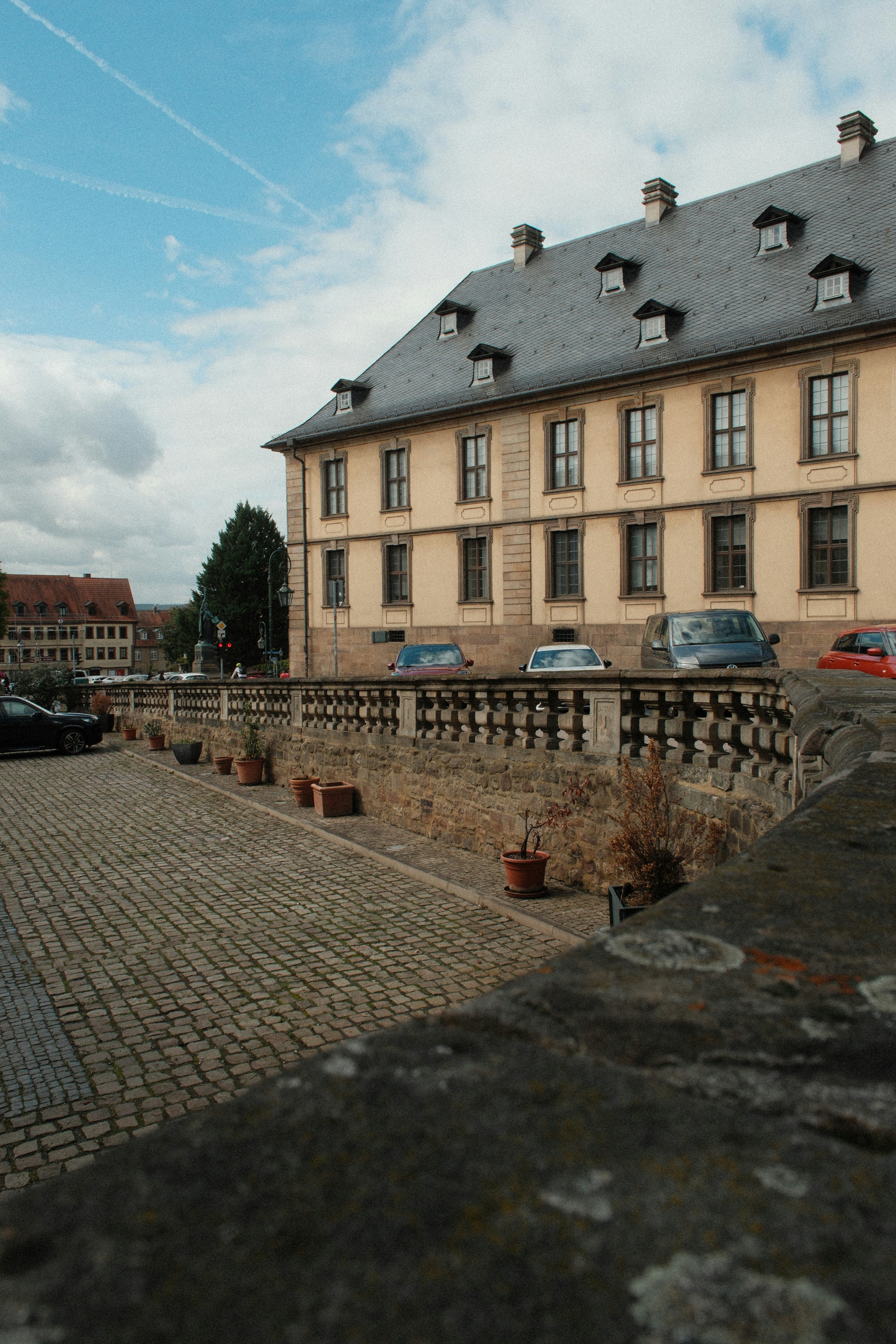 A historic yellow building with a slate roof beside a stone balustrade and cobblestone street
