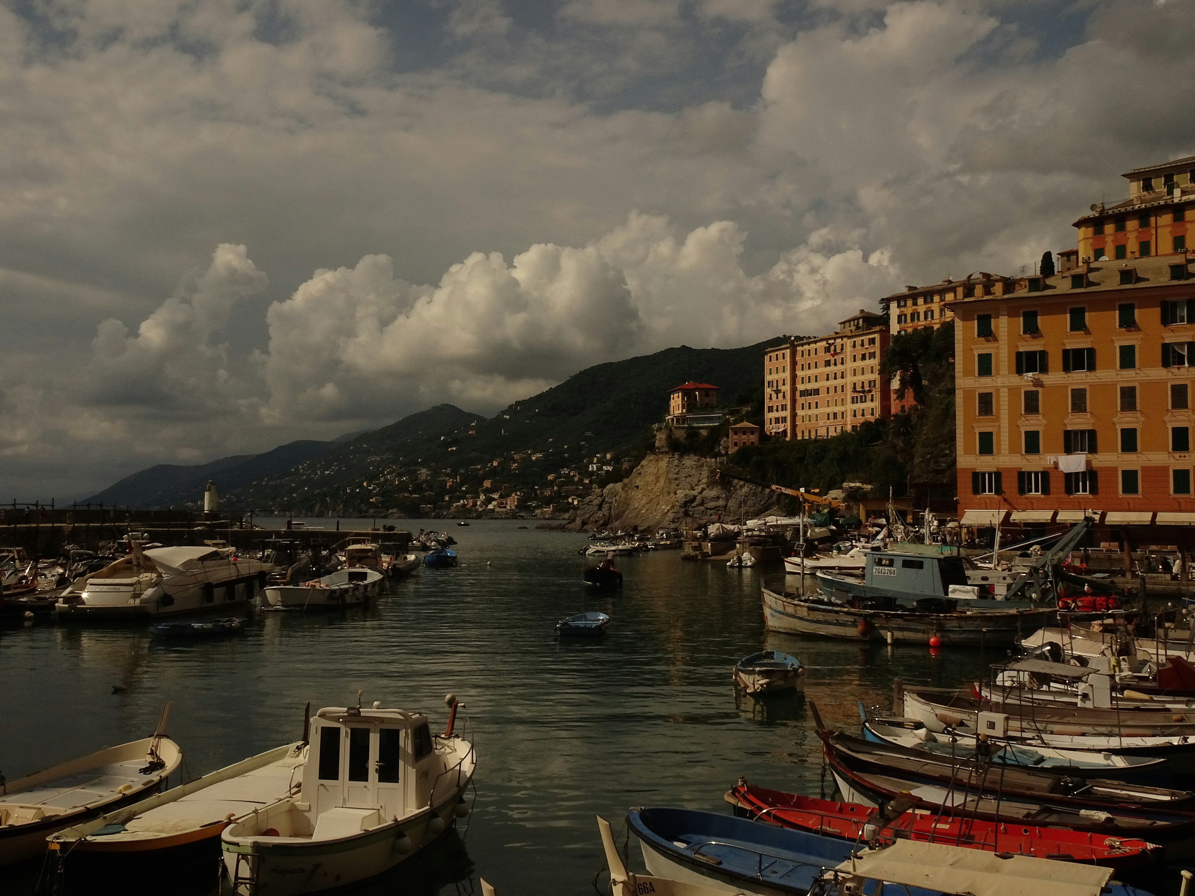 Boats moored in a harbor with yellow buildings and mountains under a cloudy sky