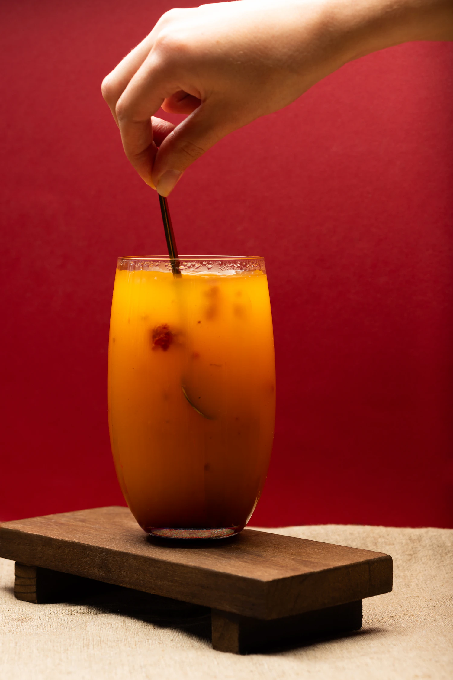 A hand stirring a blood-orange drink in a tall glass against a red wall
