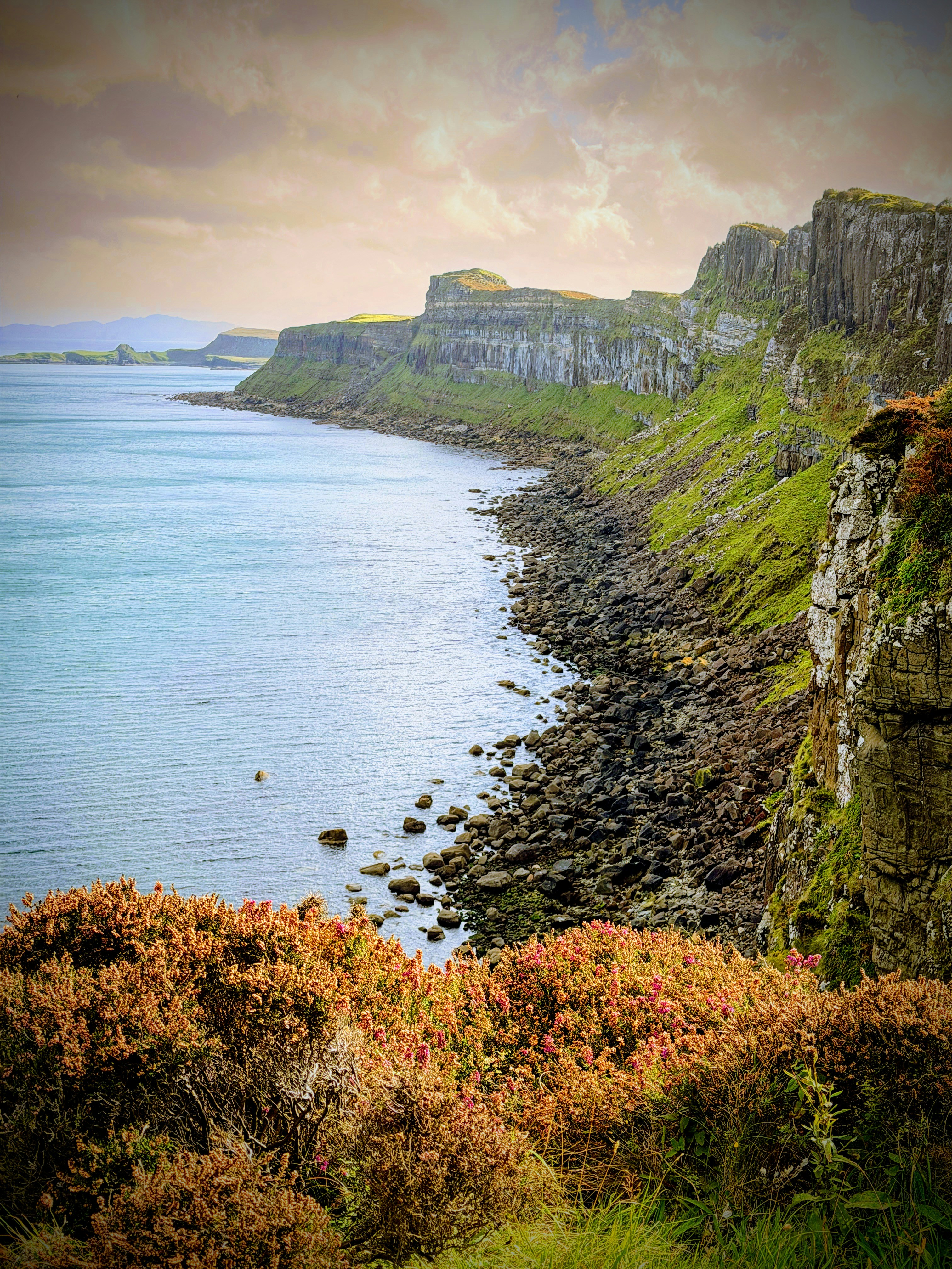 Green cliffs above a rocky shoreline and blue water under a soft sky
