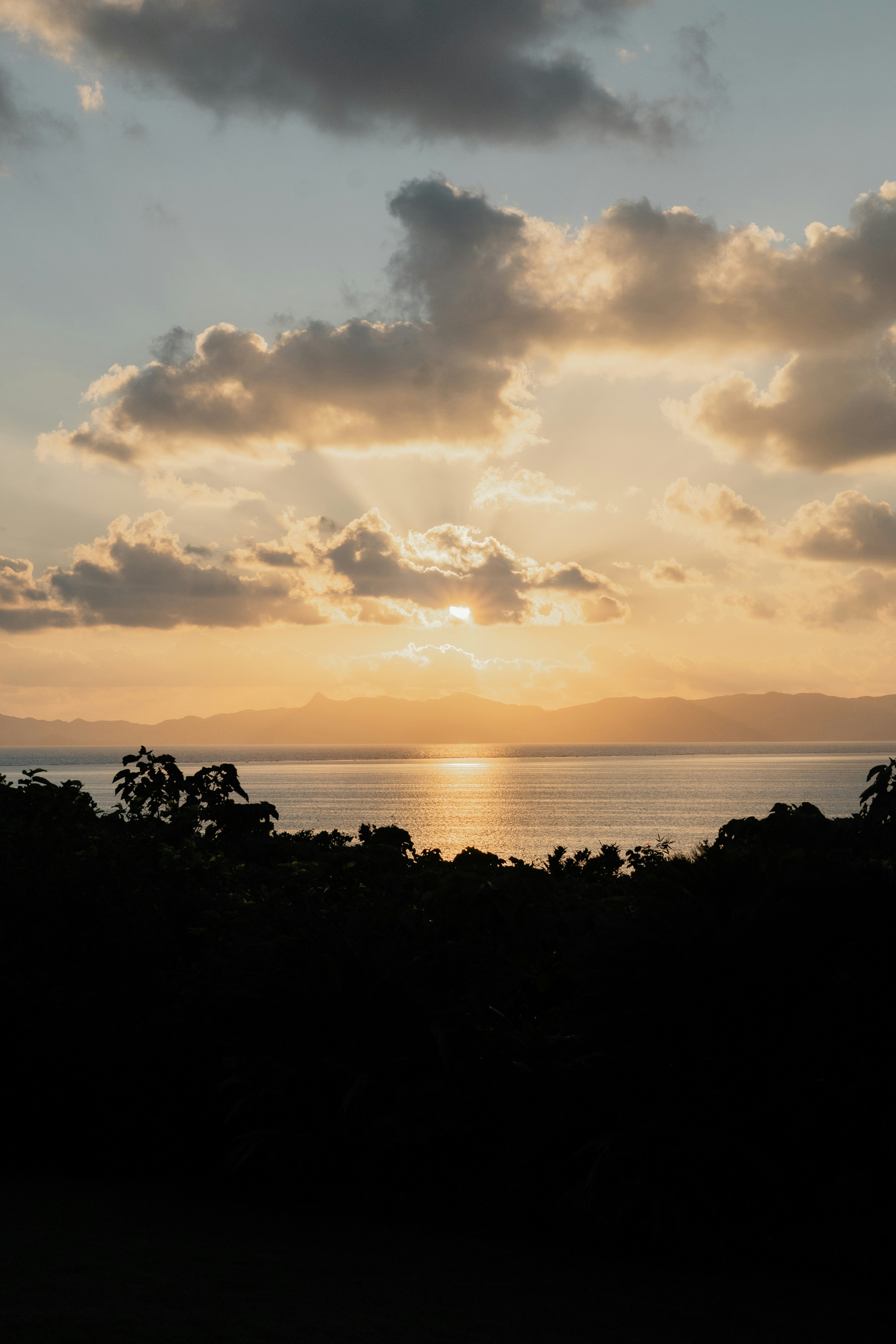Low sun glowing behind clouds over a calm sea and distant mountain silhouettes