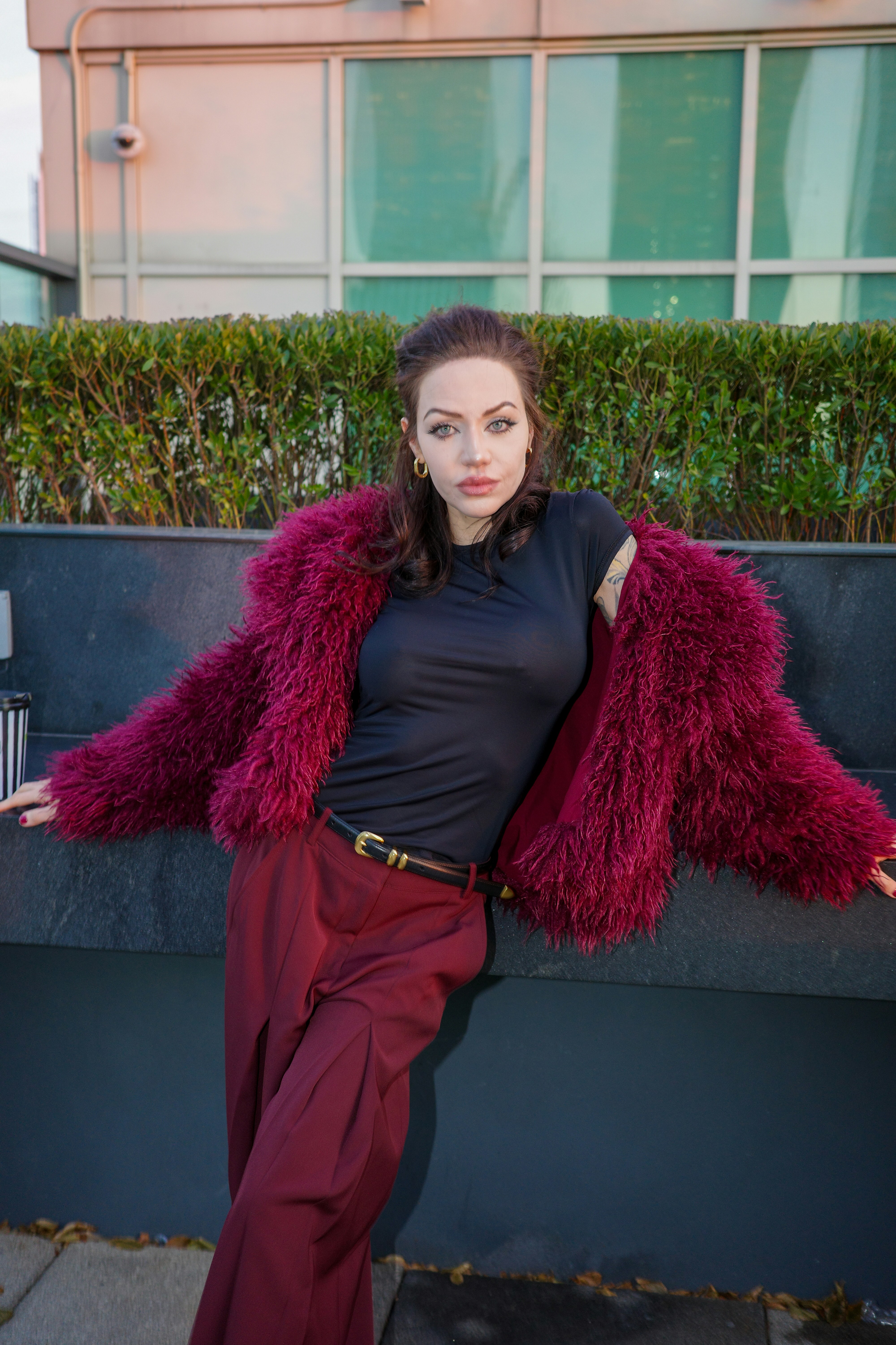 A woman in a burgundy faux fur coat and trousers leaning against a wall