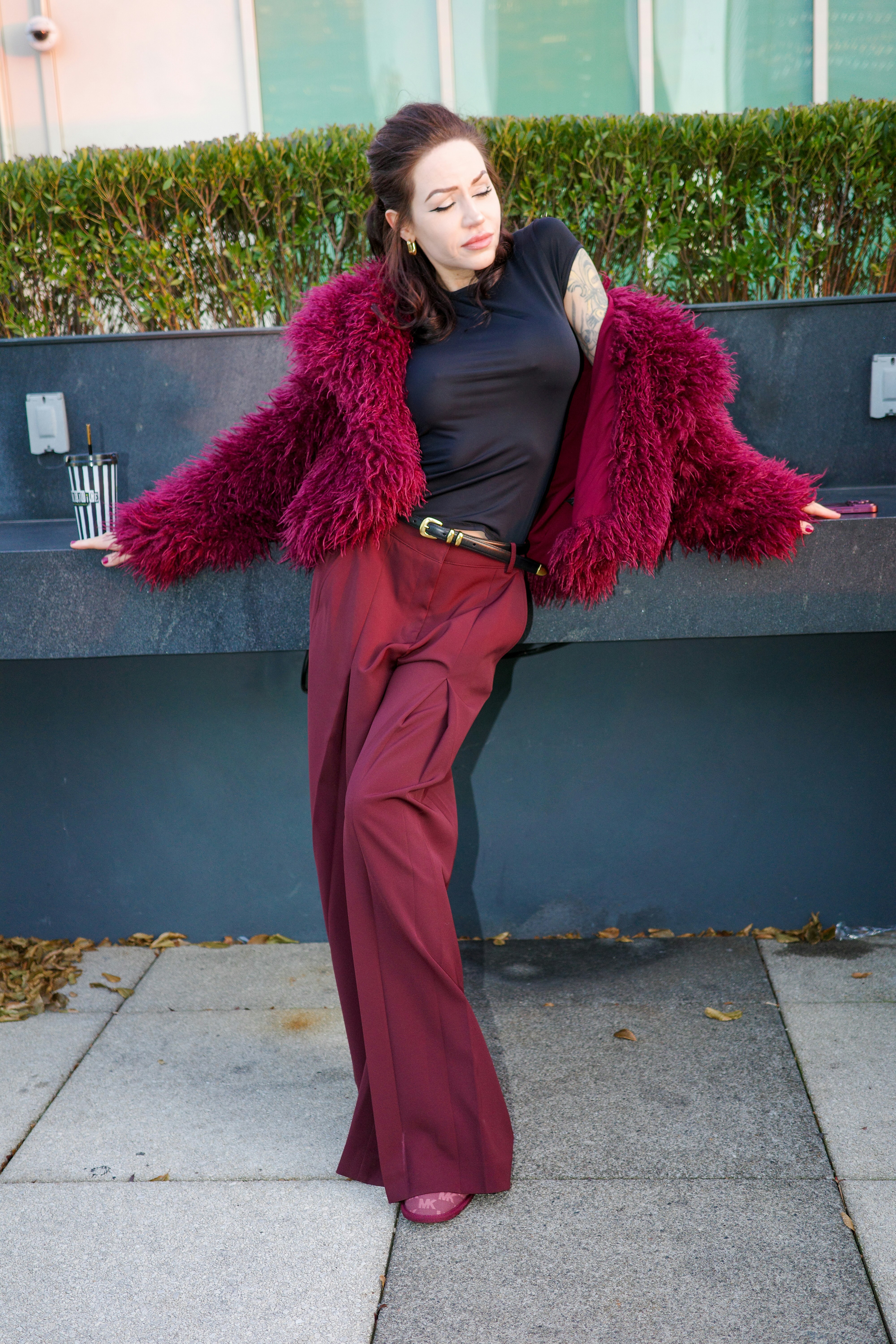 A woman in a burgundy faux fur jacket and wide-leg trousers leaning against a wall