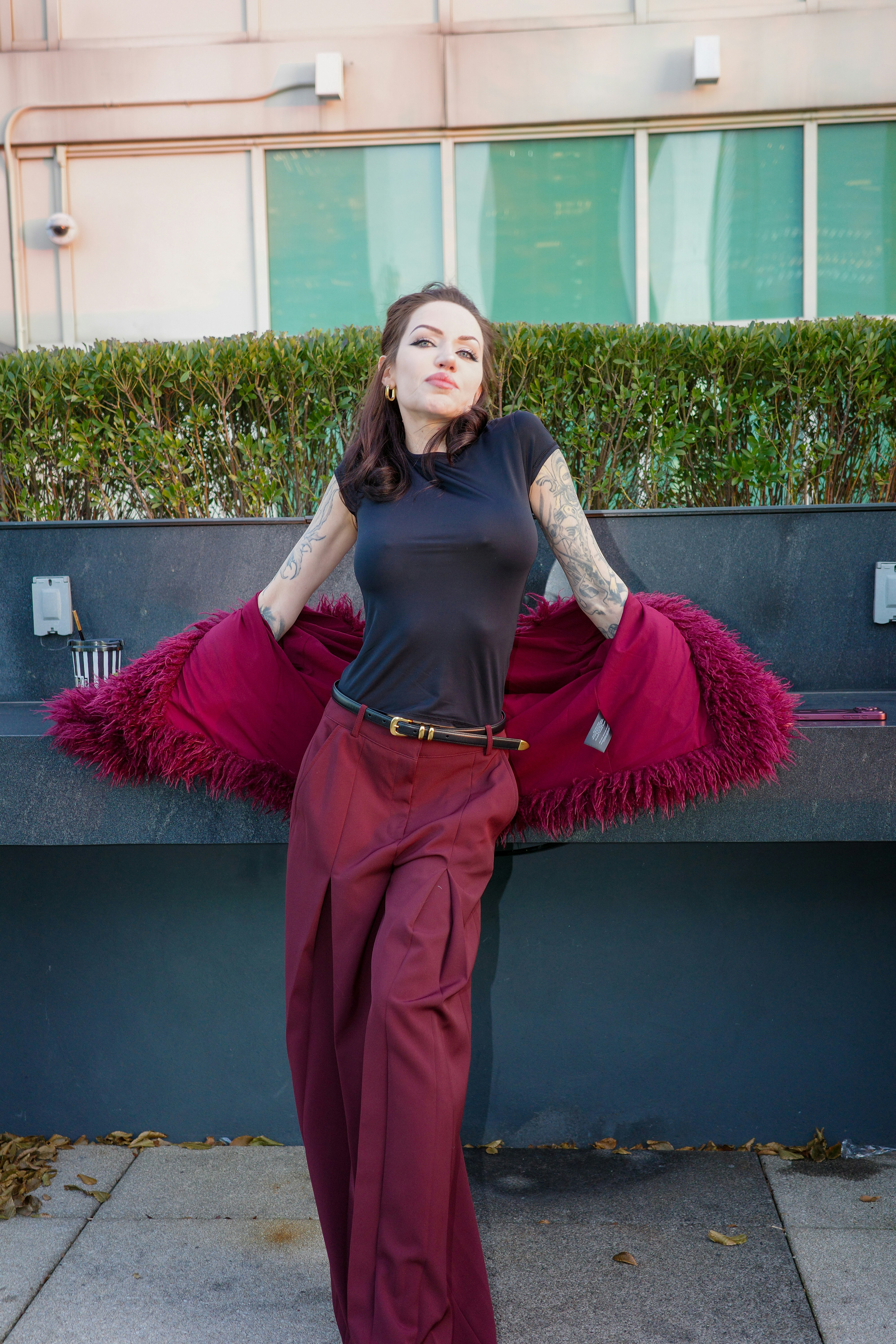 A woman in a black t-shirt and burgundy trousers leaning against a wall