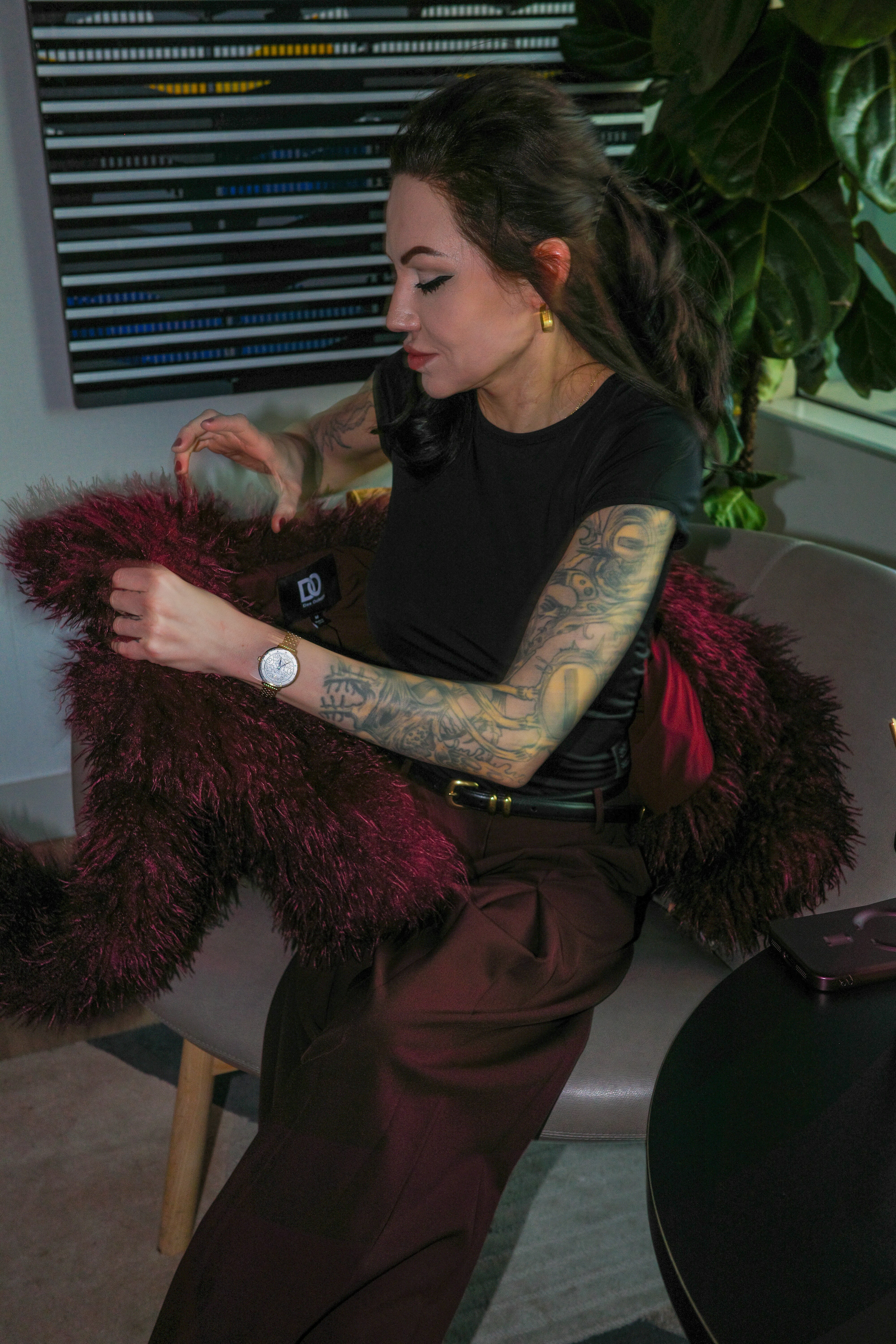 A woman with tattooed arms holding a burgundy faux fur coat in a room
