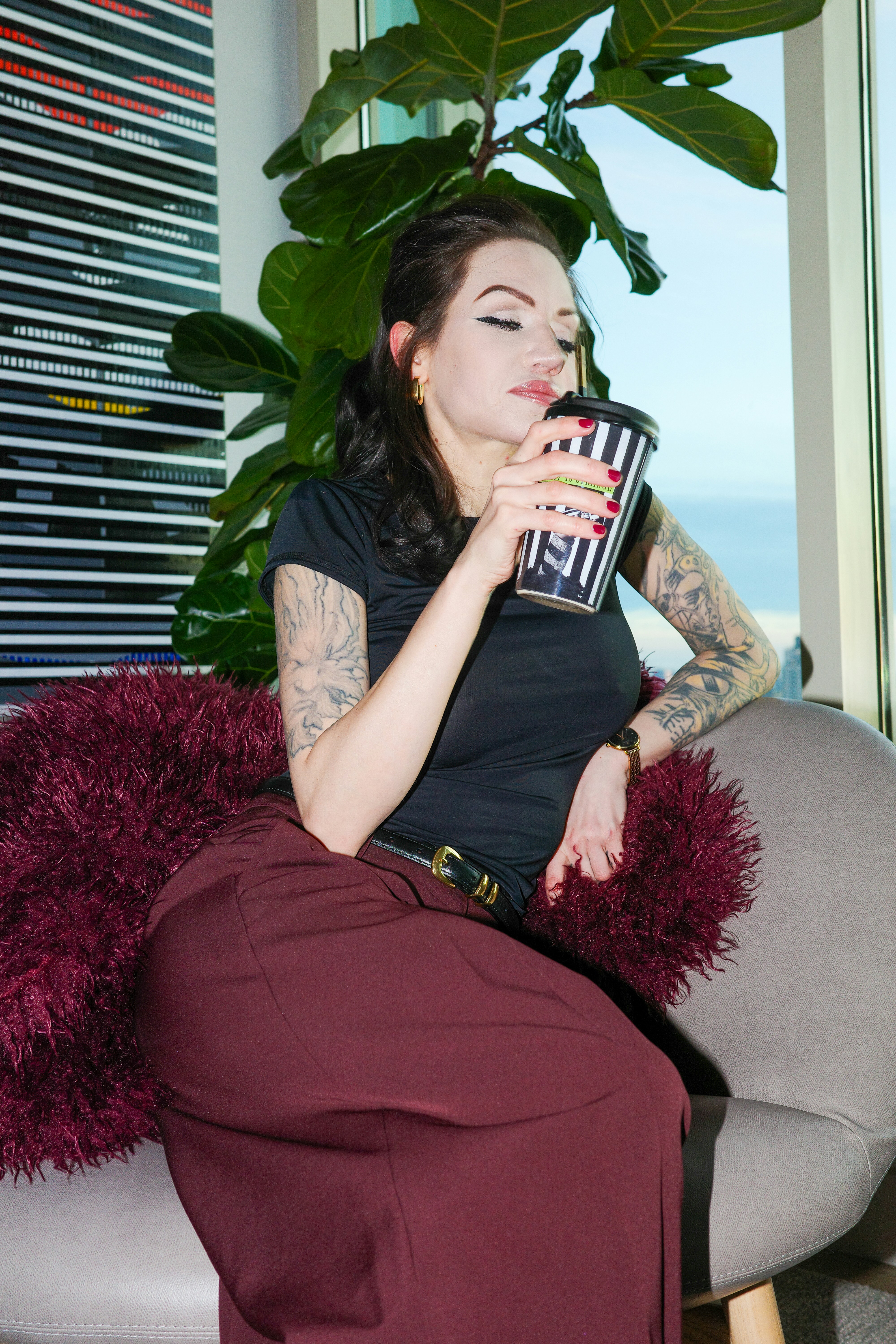 A woman with tattooed arms drinking from a striped cup in a grey chair