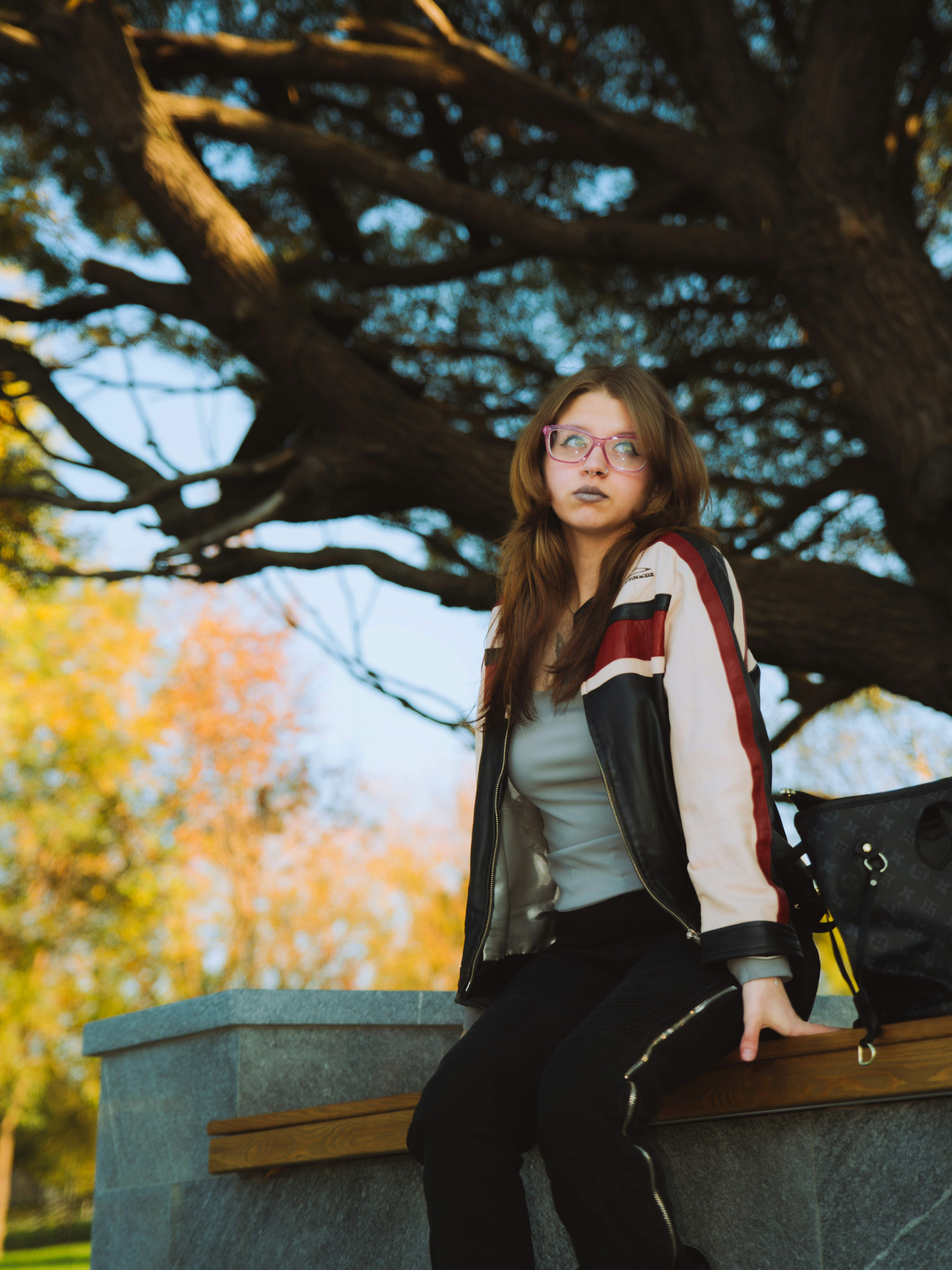 A young woman in a leather jacket and pink glasses sitting on a bench