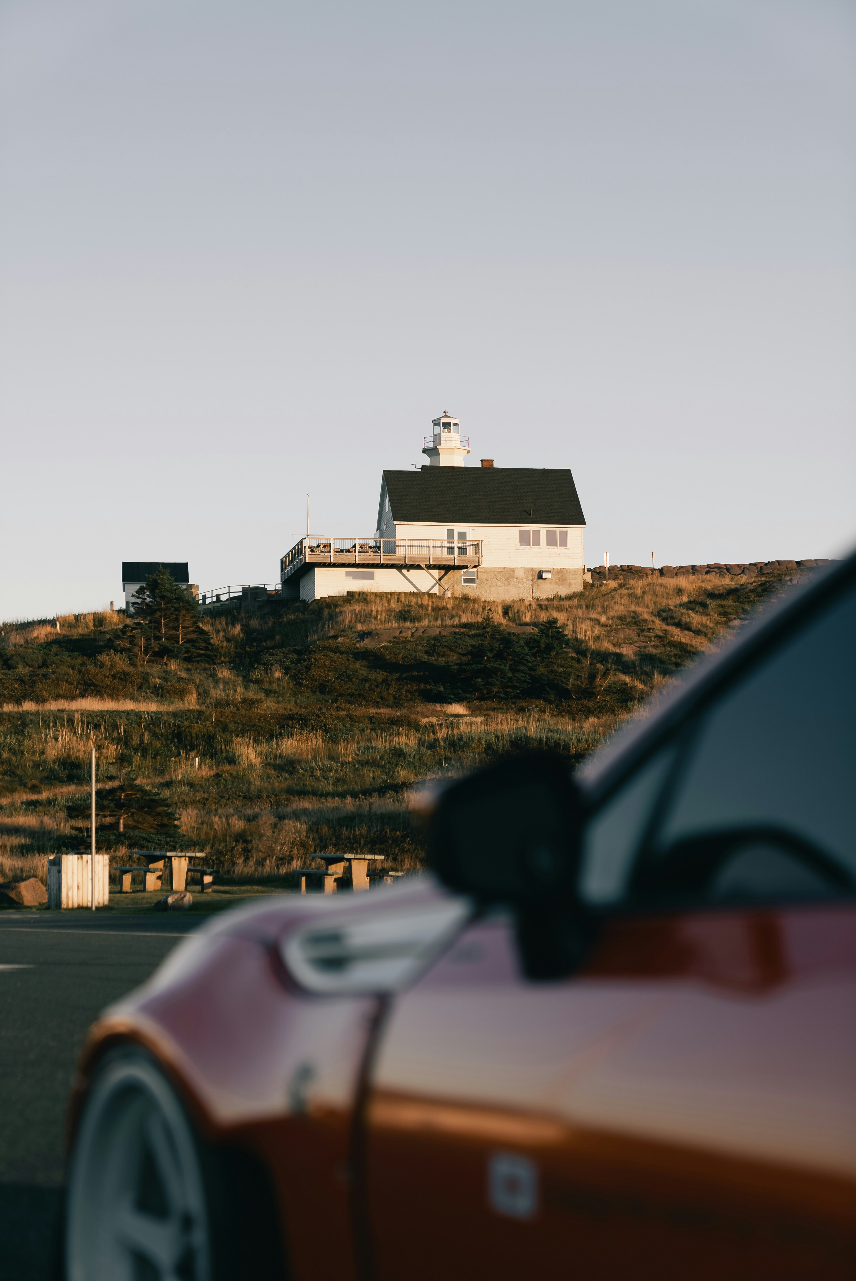 A white lighthouse on a grassy hill above a red car in the foreground