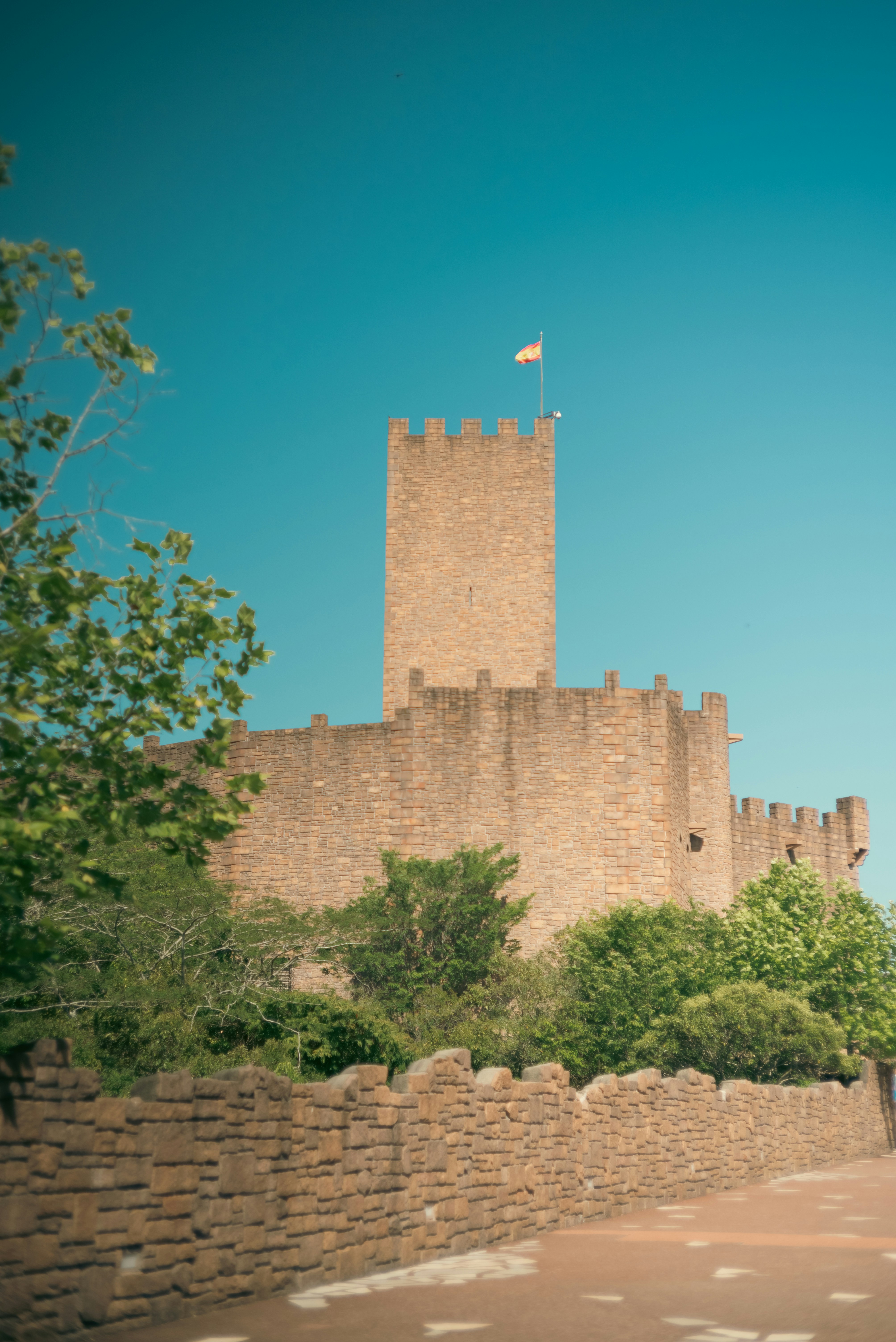 A stone castle with a tall tower and Spanish flag against a blue sky
