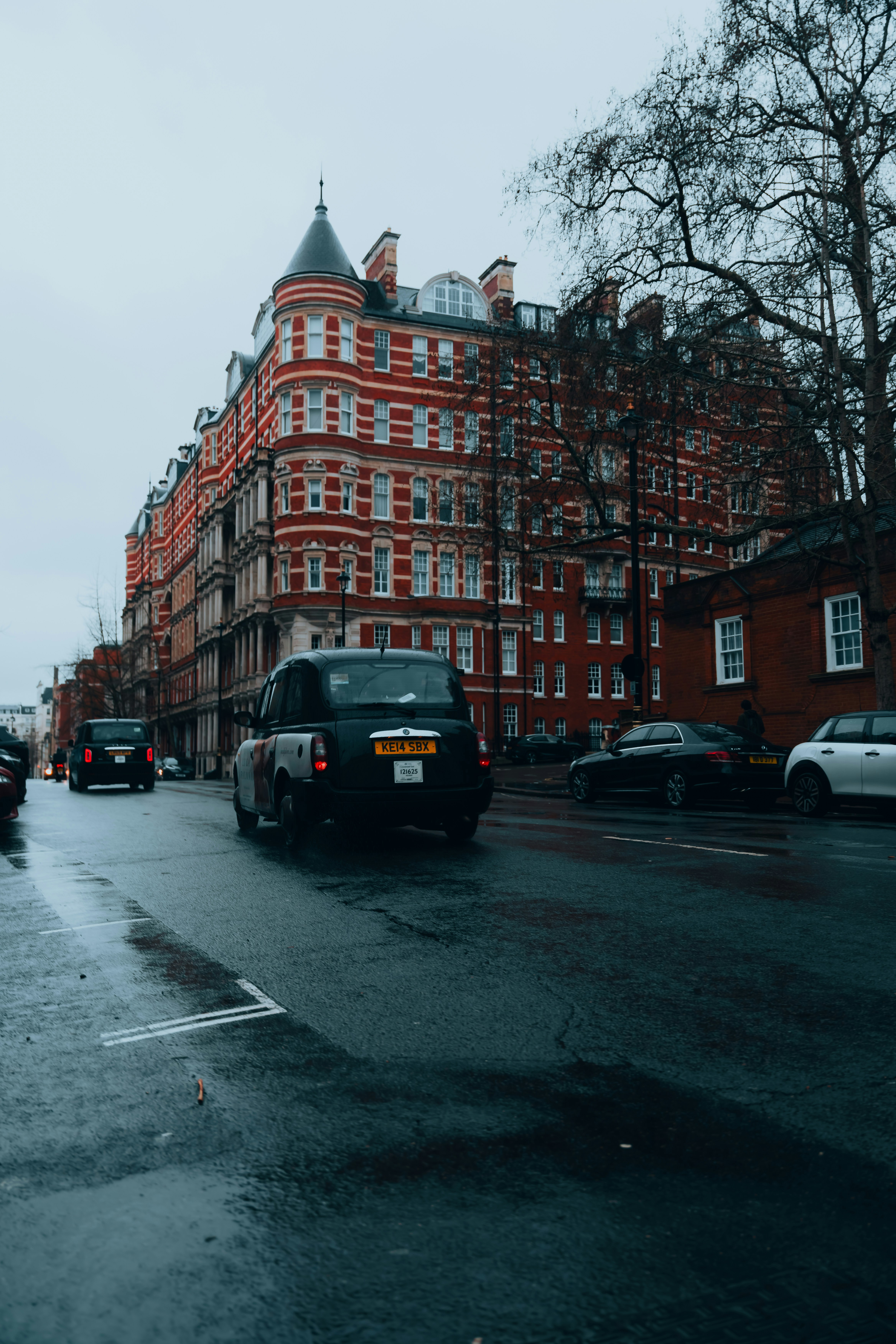 A black taxi driving on a wet street in London past red brick buildings