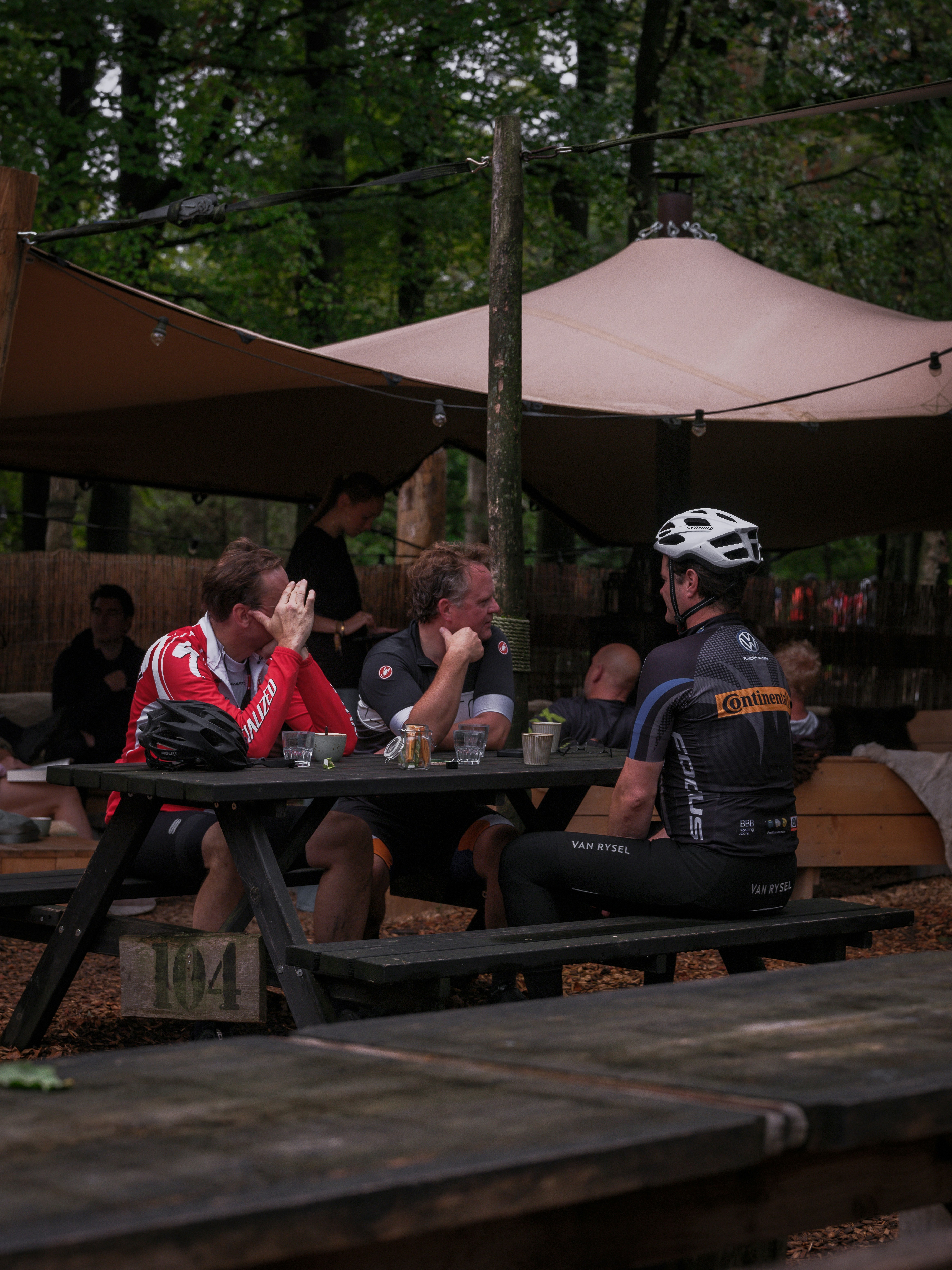 Three cyclists in athletic gear sitting at a wooden picnic table outdoors