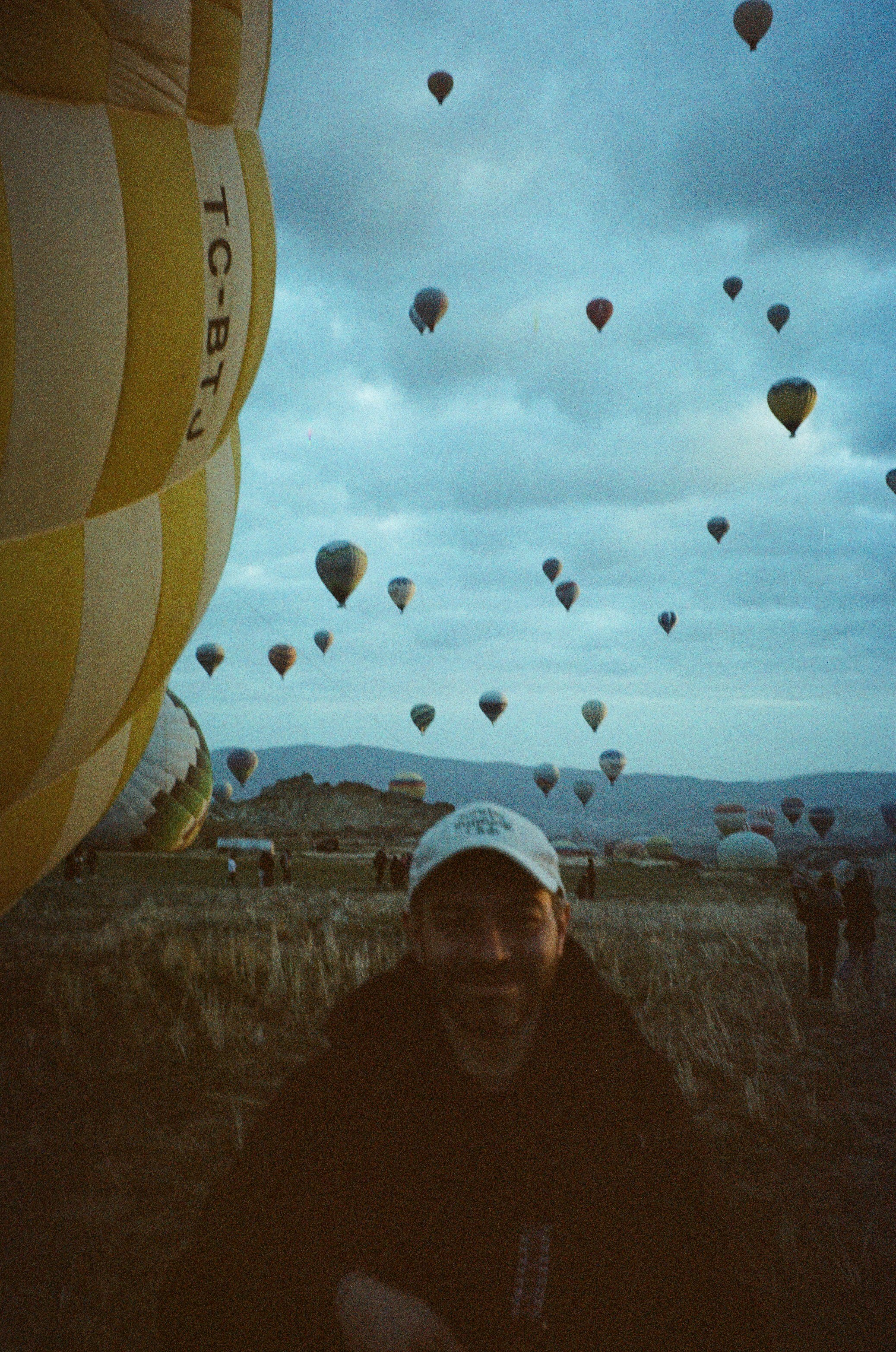 A person in a cap standing in a field with many hot air balloons