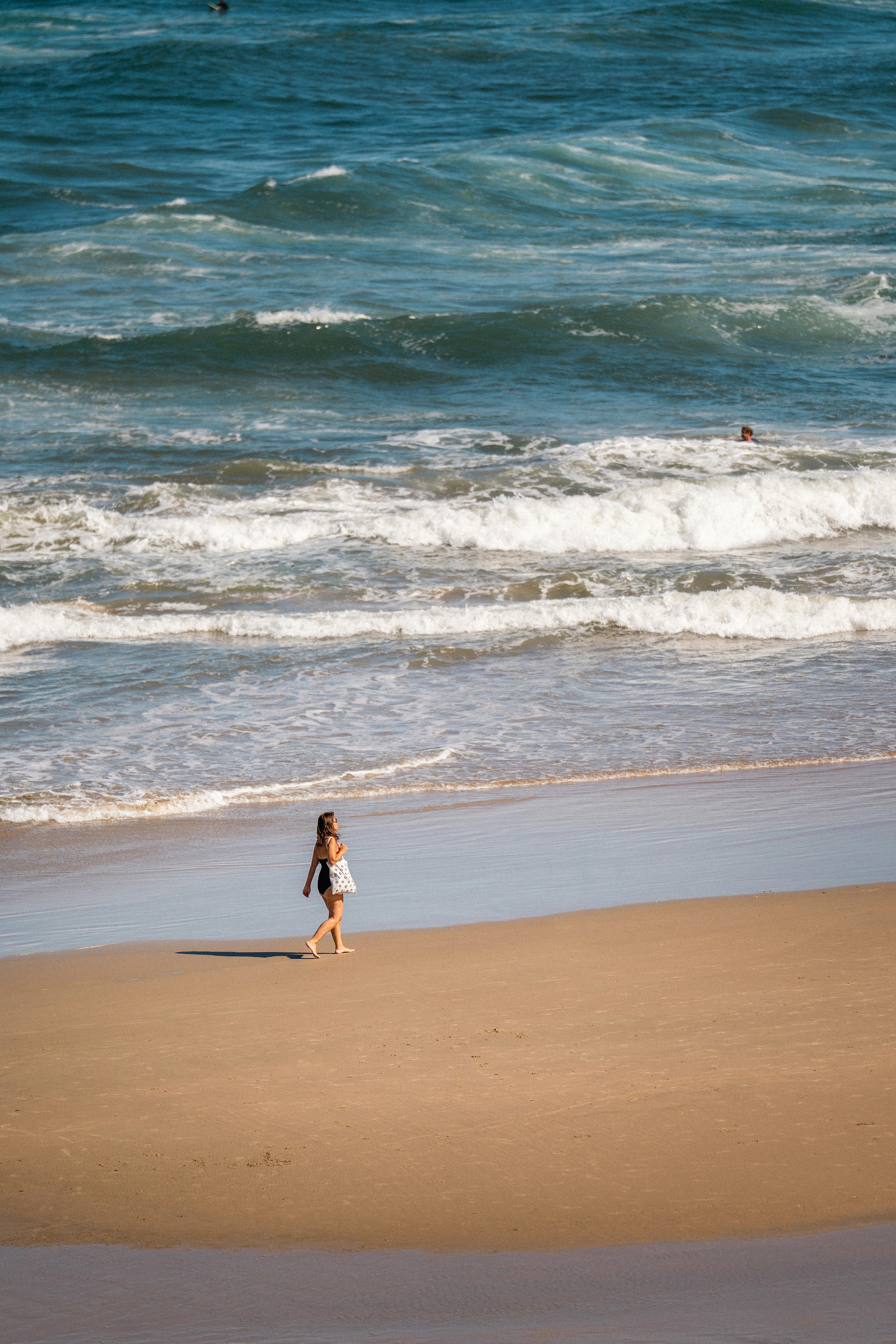 A woman in a black swimsuit walking along a sandy beach near ocean waves