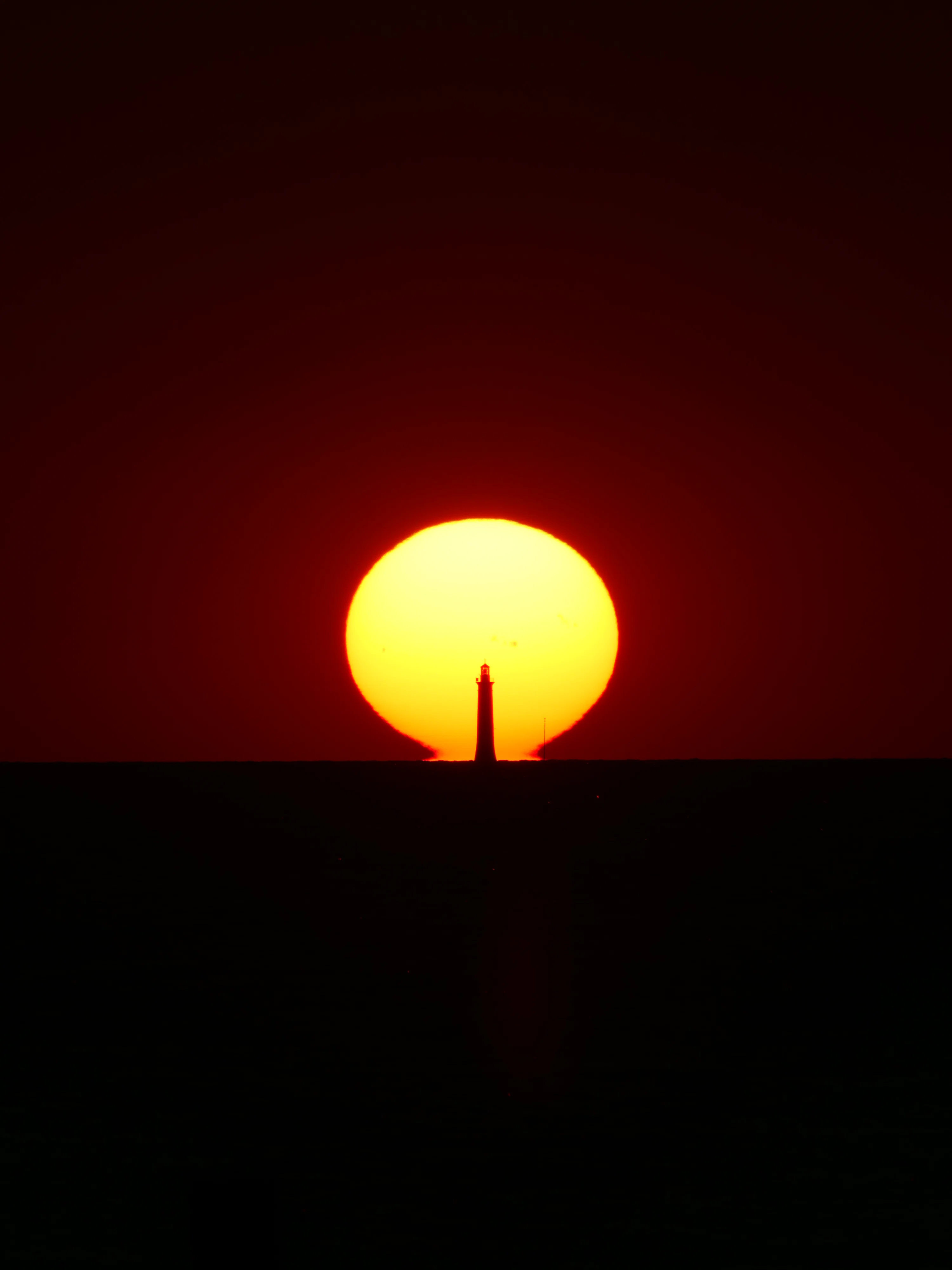 A lighthouse silhouette centered against a large low sun on the horizon