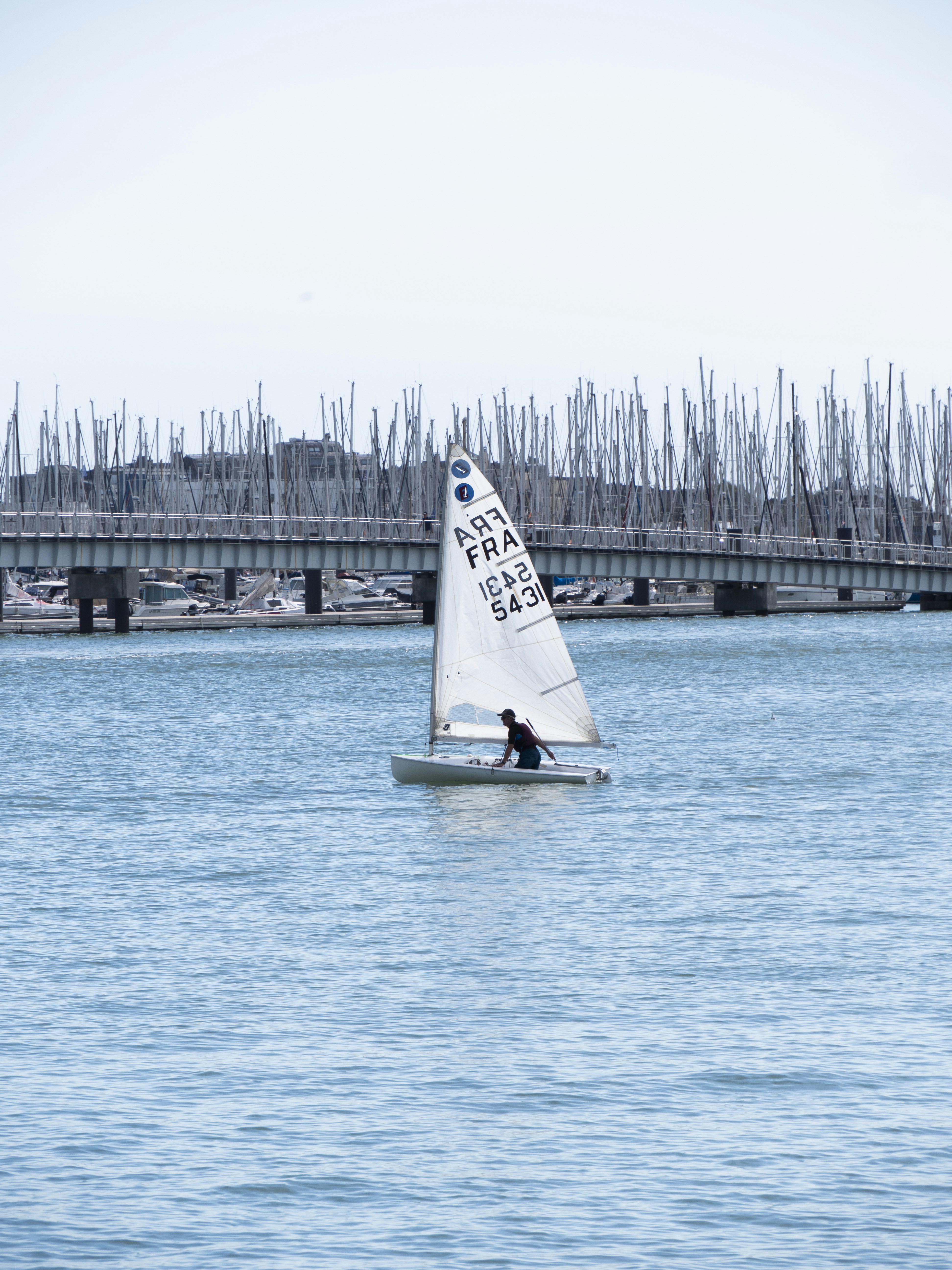 A person sailing a small white dinghy on water near a marina bridge