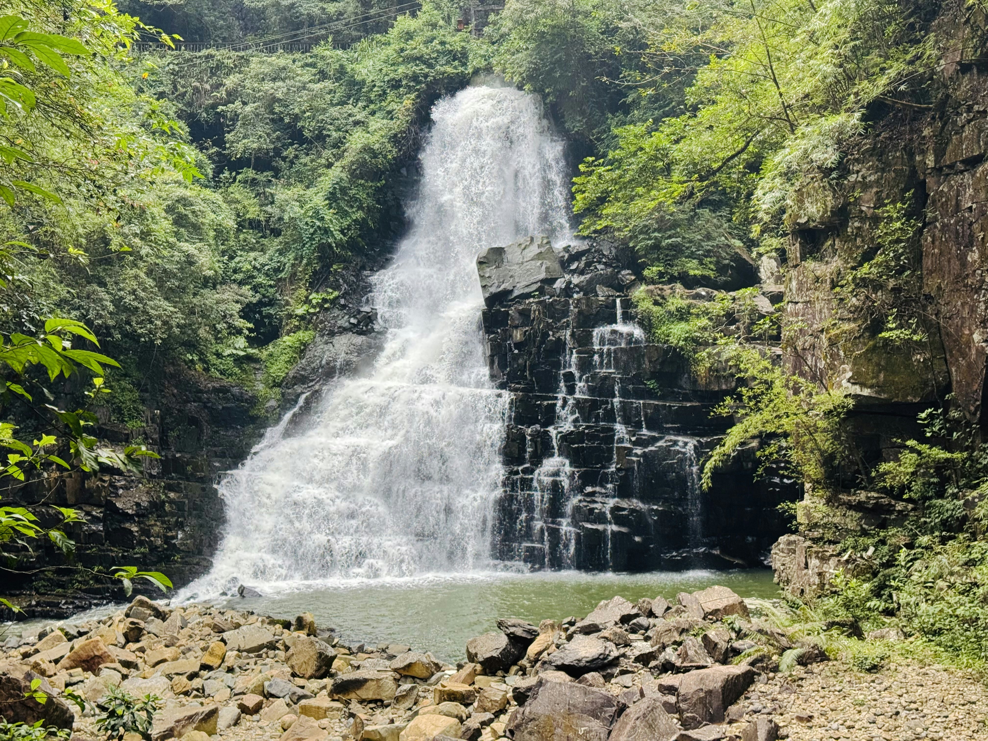 A waterfall cascading over dark tiered rocks into a pool surrounded by lush forest
